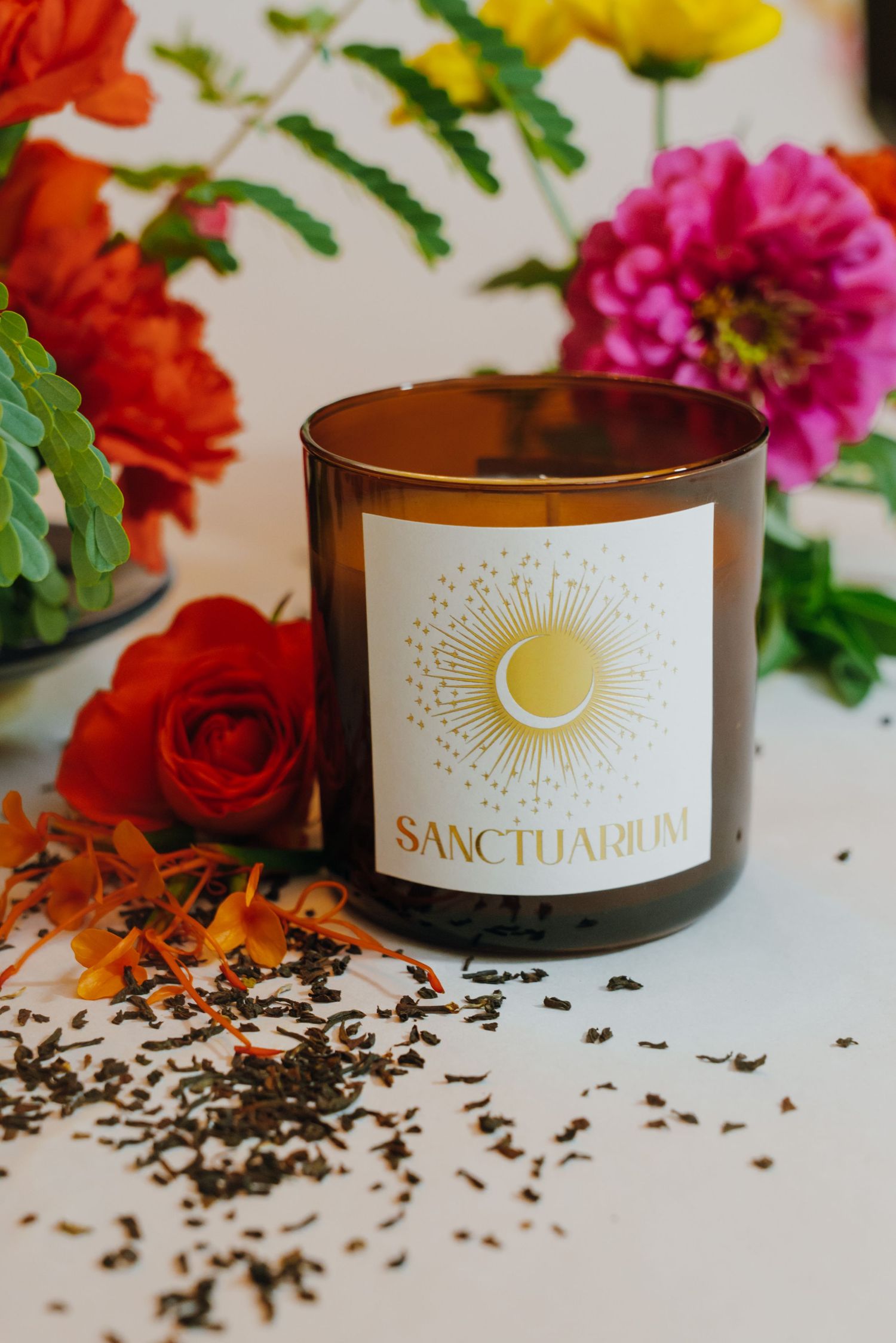 Sanctuarium amber glass candle with sun logo label surrounded by colorful flowers and loose tea leaves.