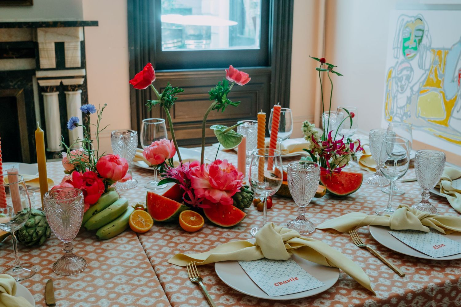 Vibrant tropical table centerpiece with pink flowers, dragon fruit, bananas, and oranges on patterned cloth.