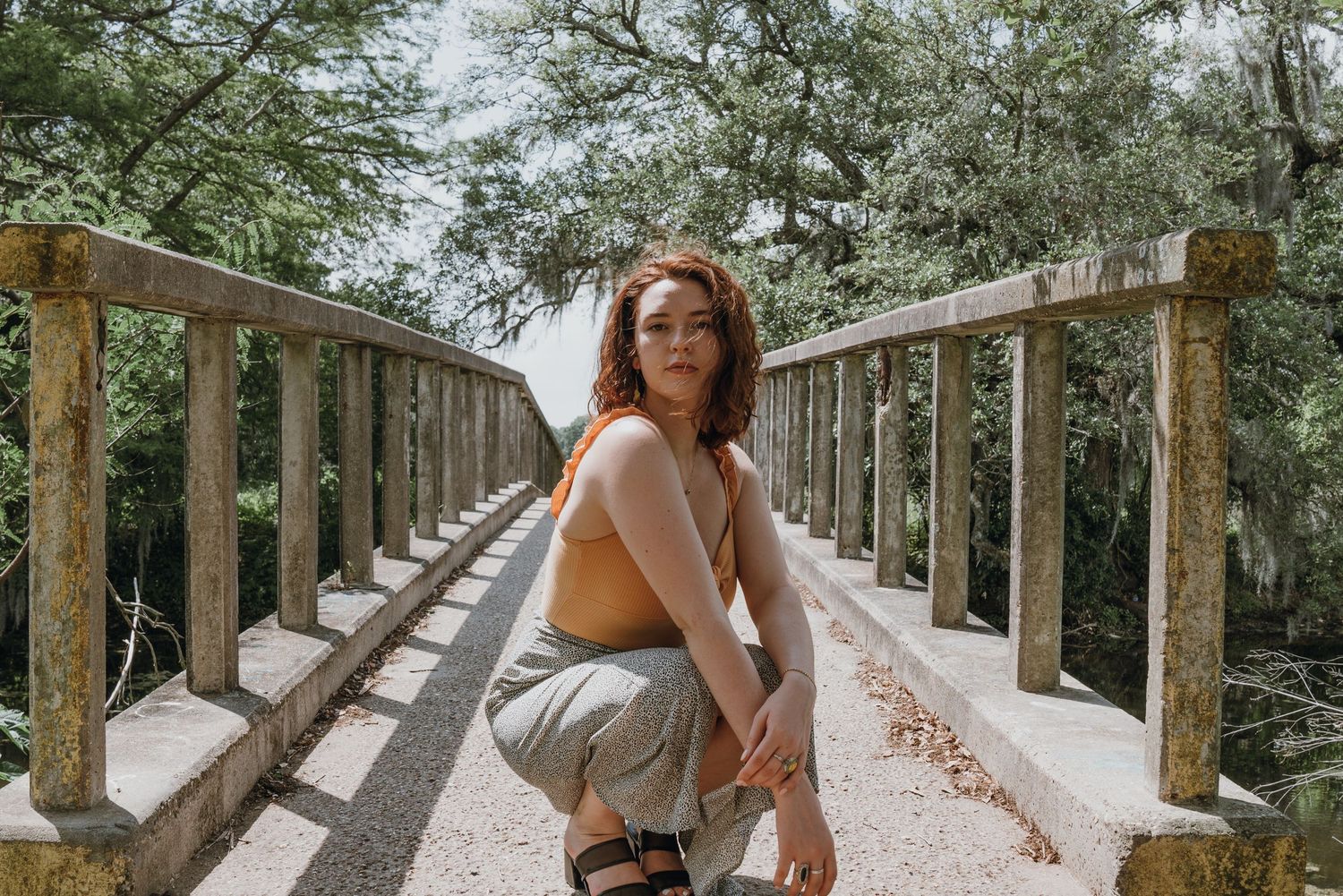 Young woman in orange top crouching on a rustic wooden bridge surrounded by lush green trees.