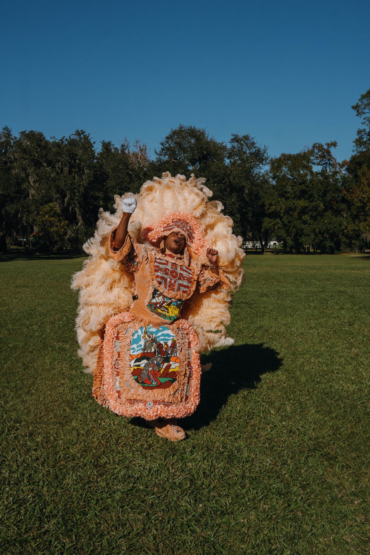 A Mardi Gras Indian in an elaborate peach feathered suit poses in a sunny park.