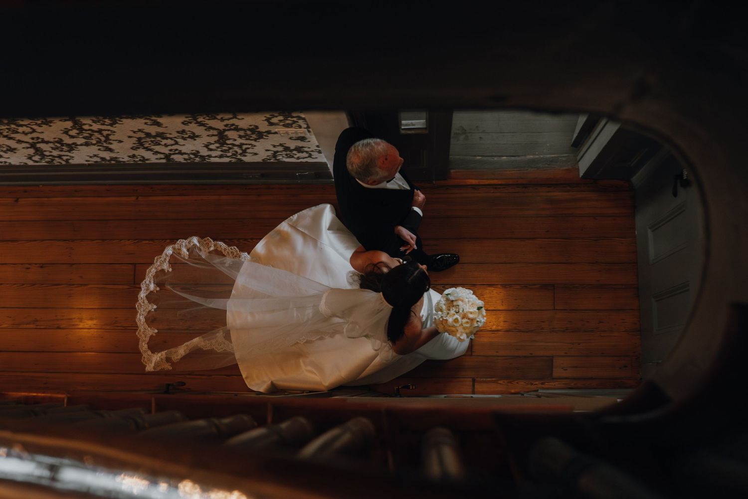 Aerial view of bride in white gown sitting on wooden staircase holding a bouquet.