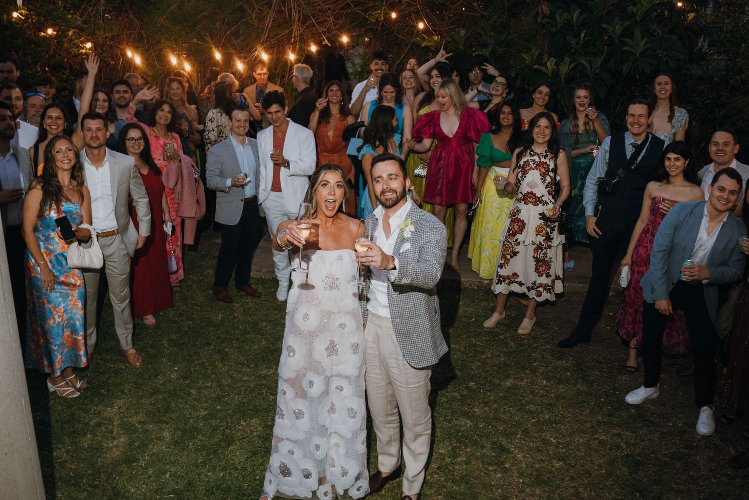 Bride and groom celebrate with sparklers surrounded by cheering wedding guests at outdoor reception.
