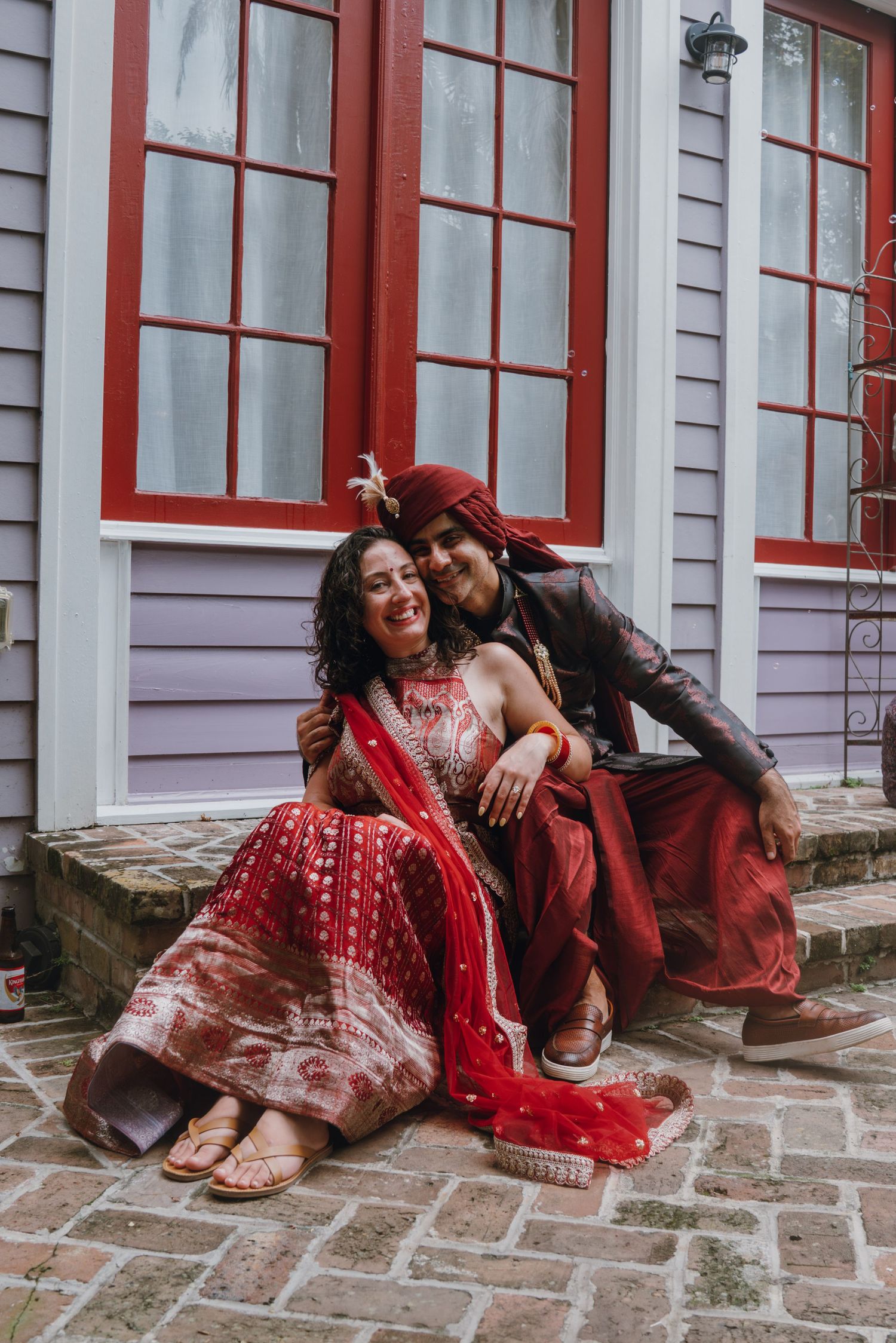 Indian bride and groom sitting together on brick steps smiling in traditional red wedding attire.