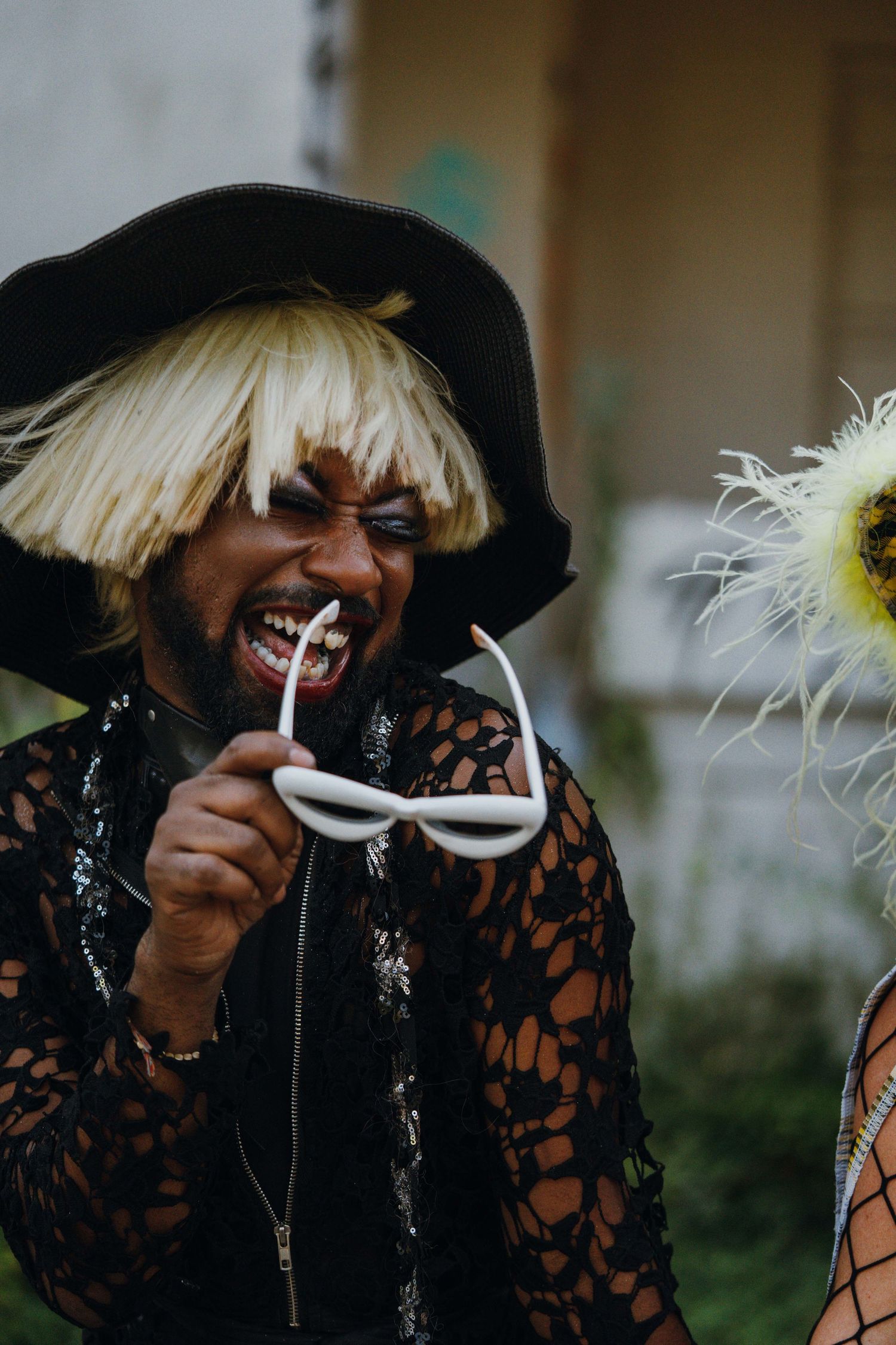 Person in blonde wig and black hat biting white sunglasses at a colorful costume party outdoors.