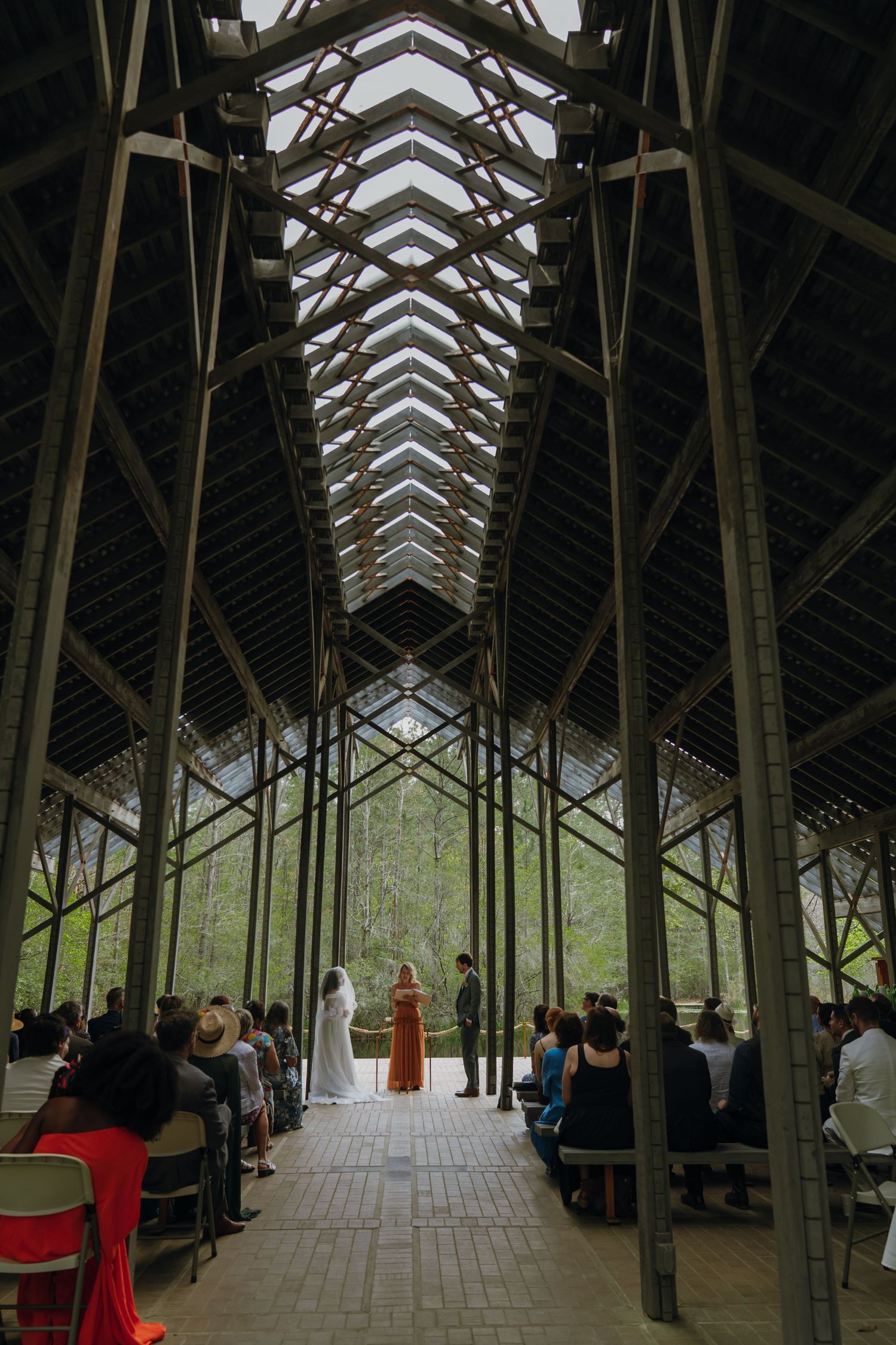 Wedding ceremony inside stunning glass and steel chapel surrounded by forest with guests seated in rows.