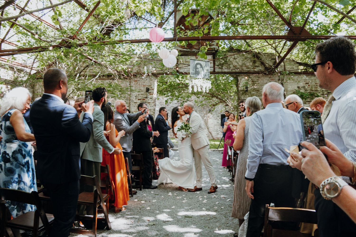 Bride and groom walk back down aisle as guests celebrate at outdoor garden wedding ceremony.