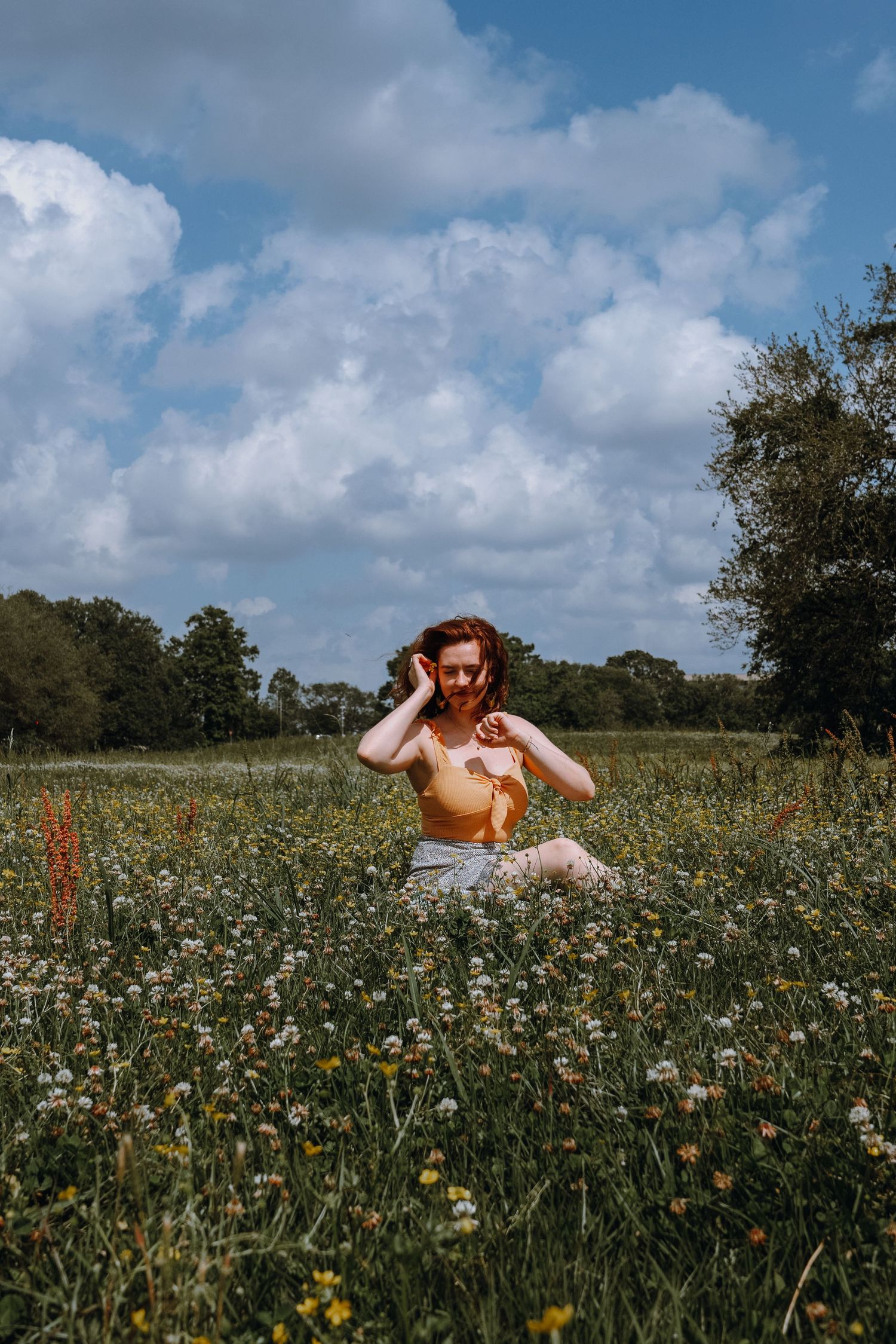 Redheaded woman in yellow top sitting in a lush wildflower field under a dramatic cloudy sky.