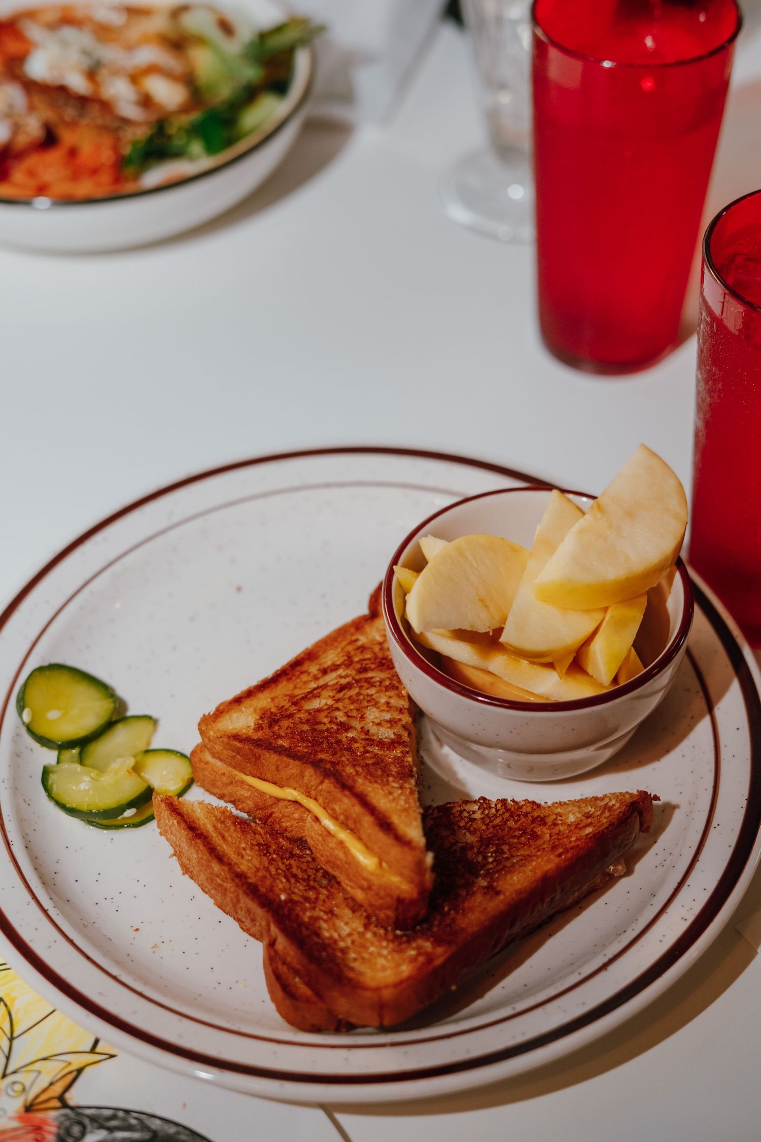 Grilled cheese sandwich with apple slices and pickle chips on vintage diner plate with red glass.