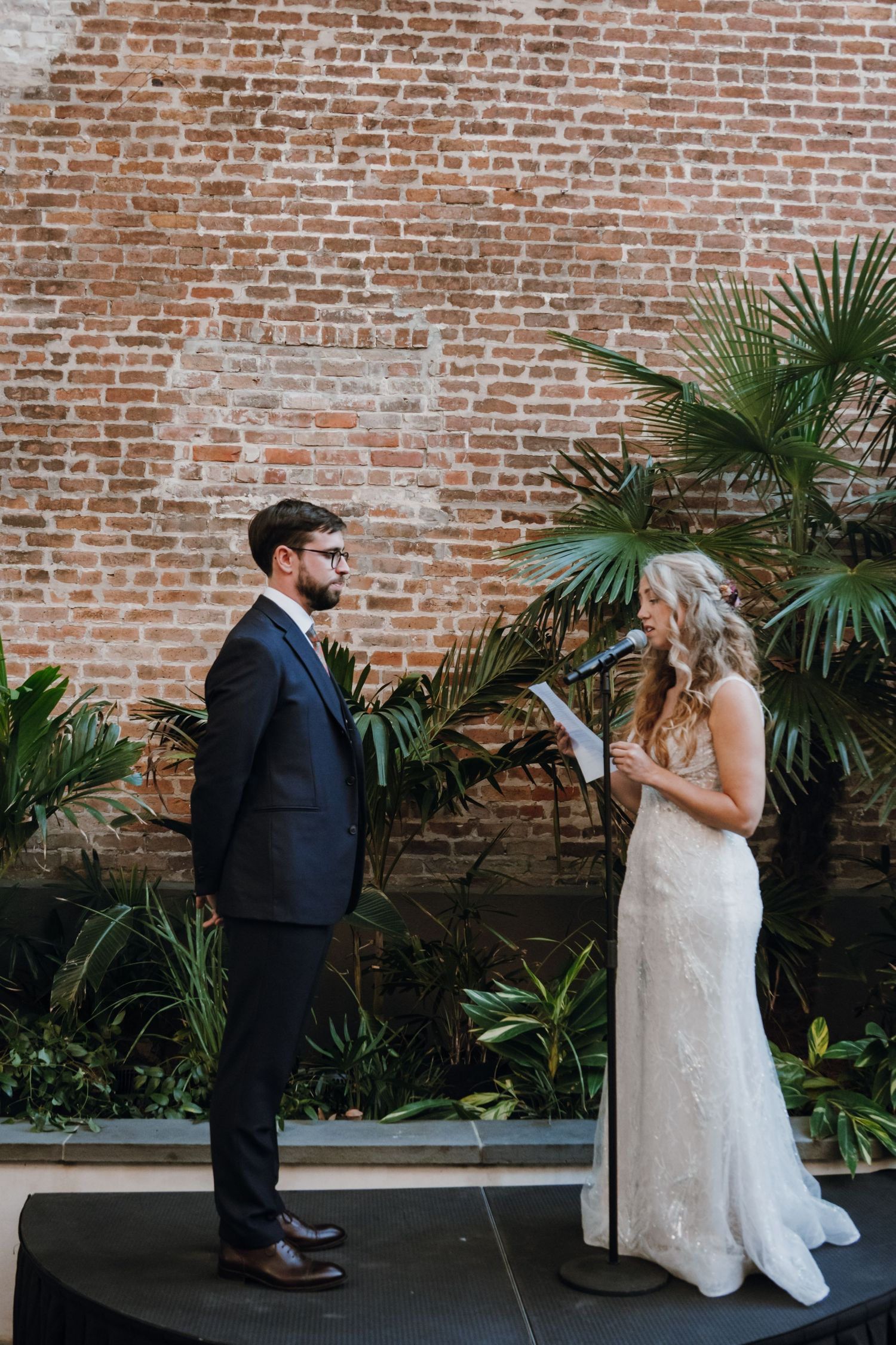 Bride reads wedding vows from paper to groom in navy suit against exposed brick wall with palms.