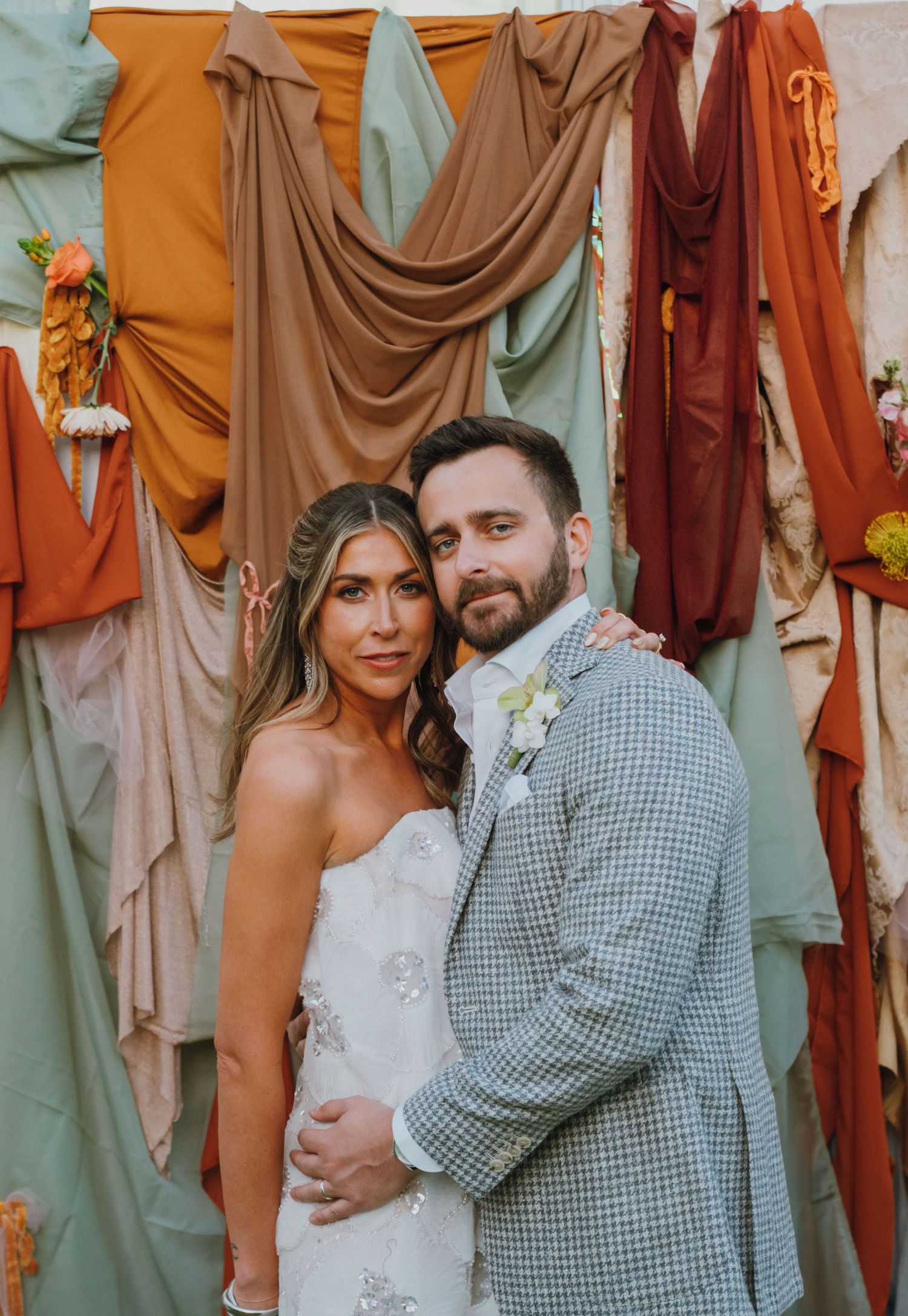 Bride and groom pose together in front of colorful draped fabric backdrop at boho wedding reception.