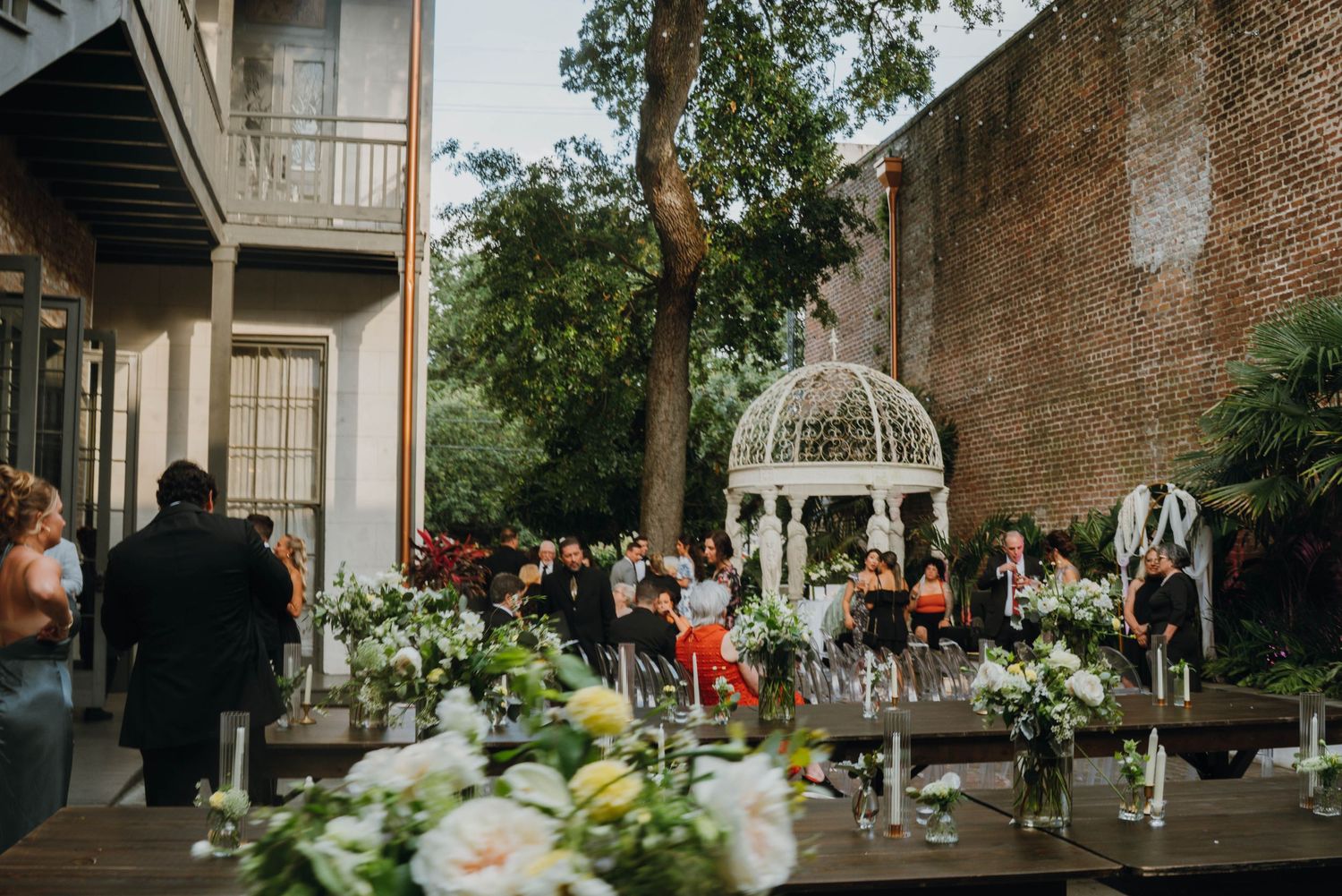 Outdoor wedding reception in a courtyard with floral arrangements, gazebo, and brick wall backdrop.