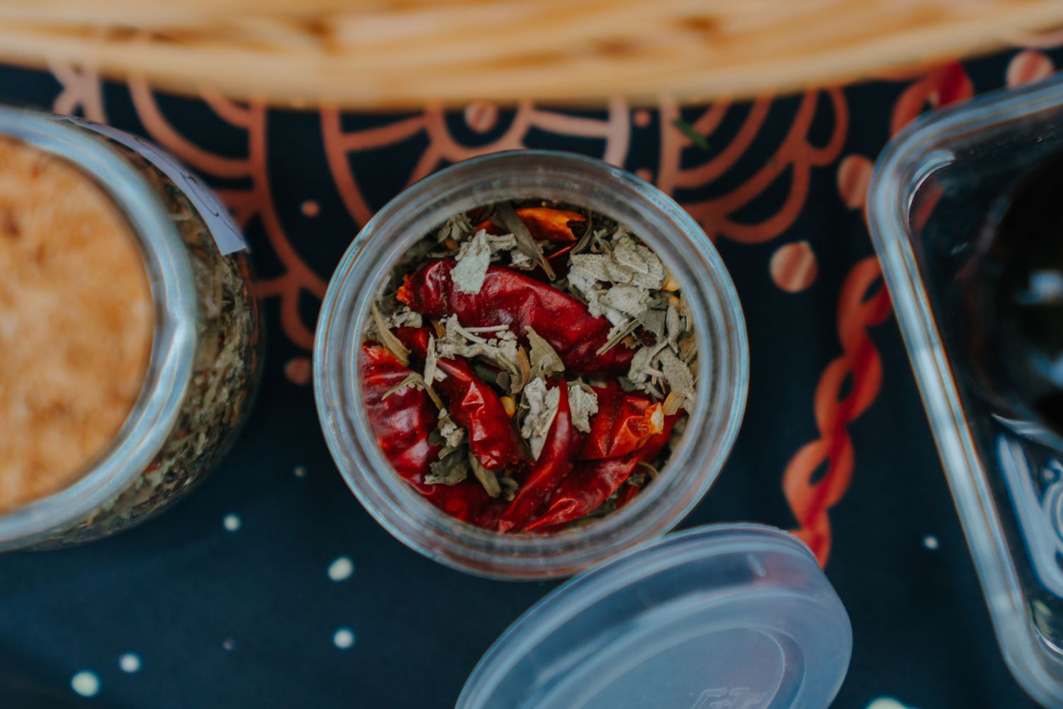 Glass jar filled with dried red chili peppers and mixed herbs, surrounded by food containers on a dark surface.