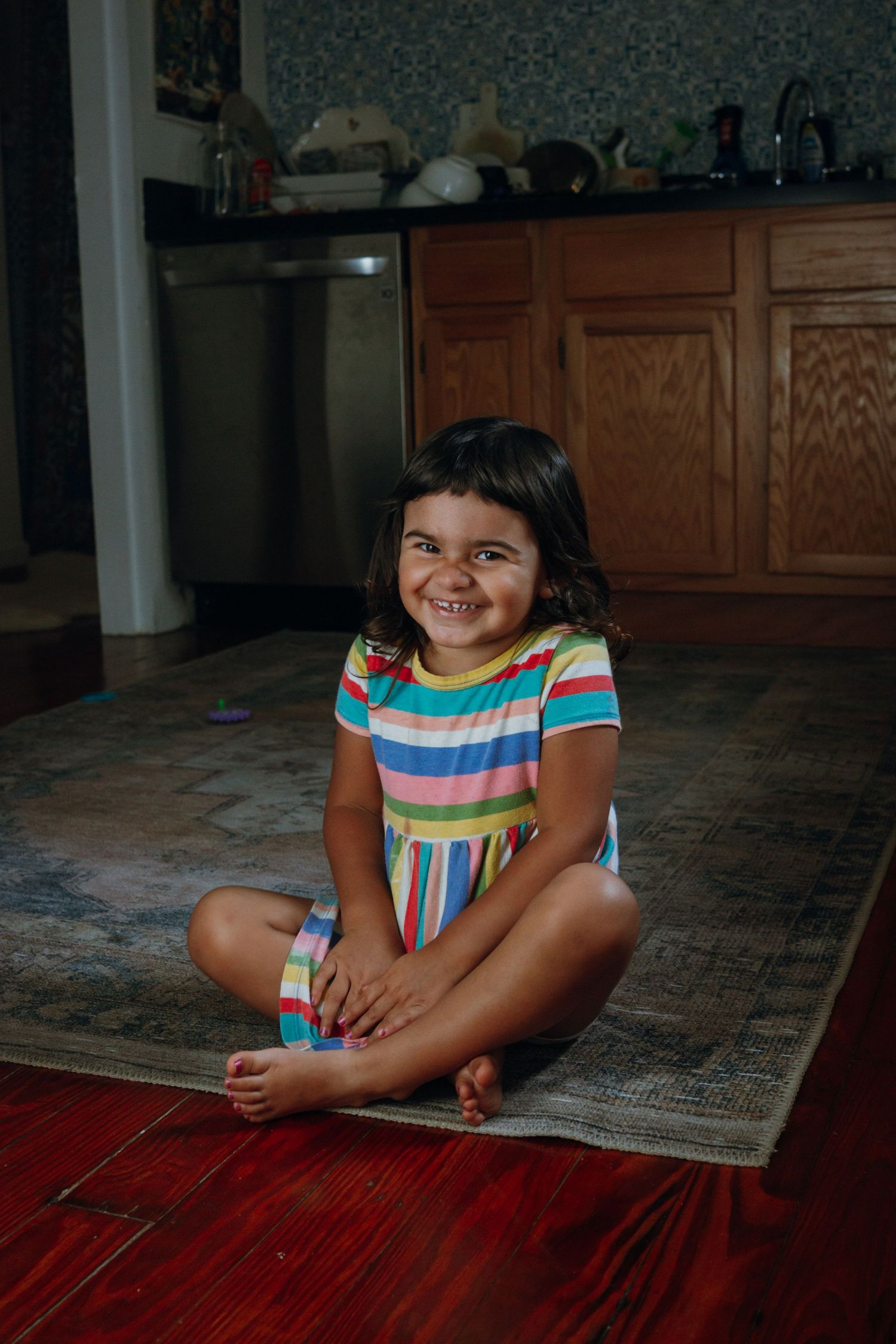 Young girl in colorful striped dress sitting cross-legged on rug, smiling happily in a home kitchen setting.