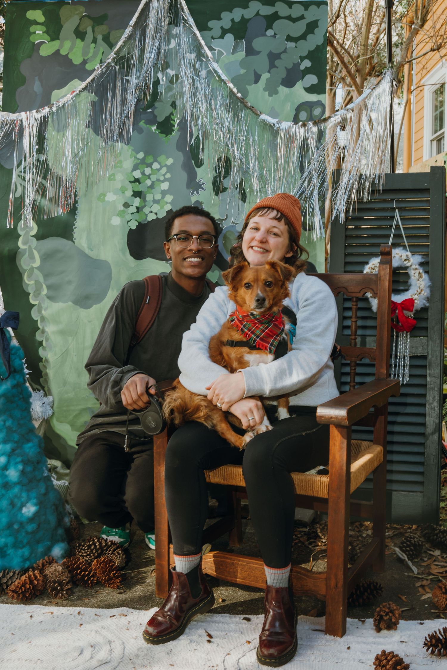 Couple sitting on wooden bench with brown dachshund wearing plaid bandana, posing before holiday mural with pine cones.