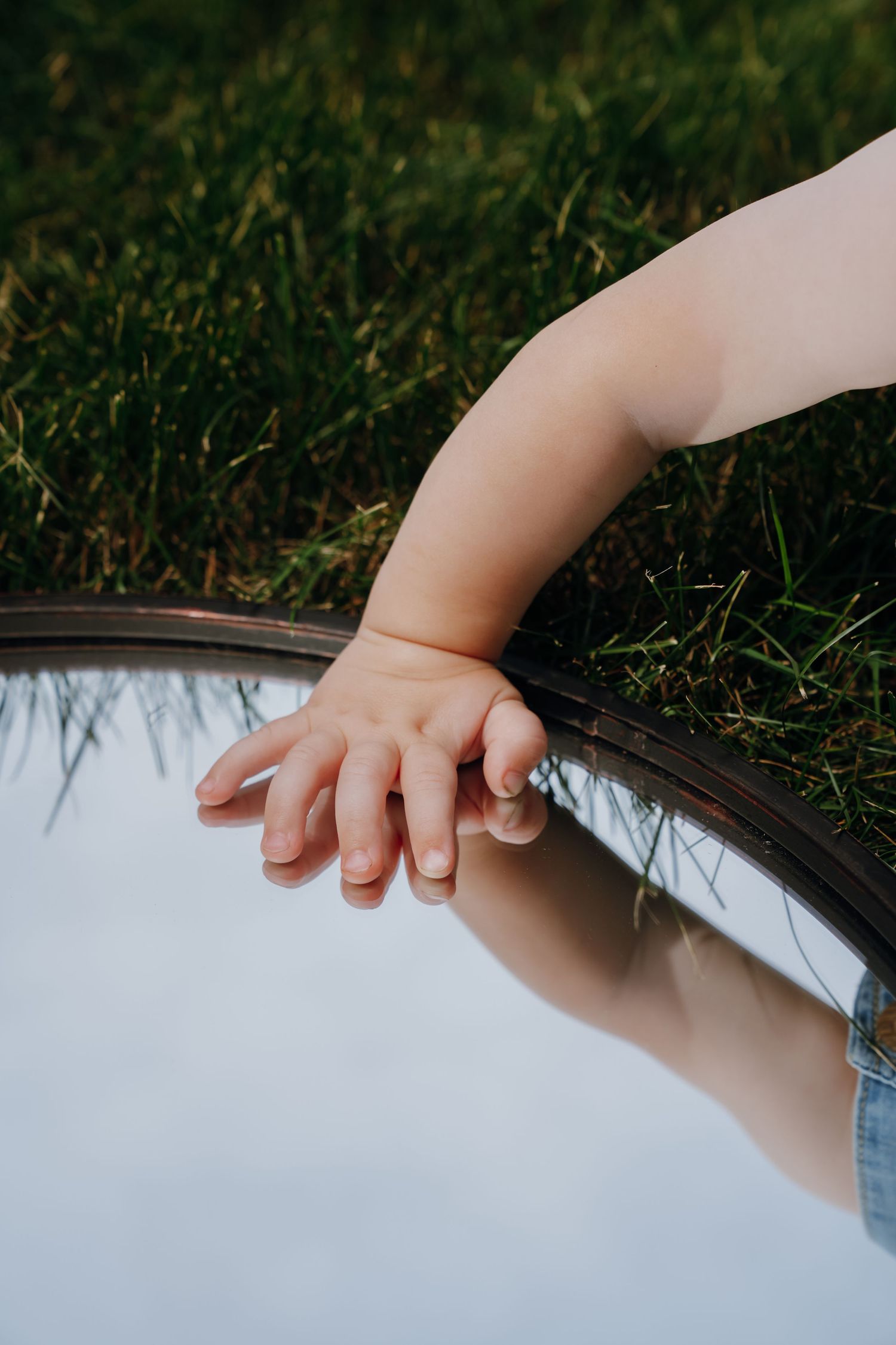 A baby's hand rests on the edge of a round mirror reflecting the sky, placed on green grass.