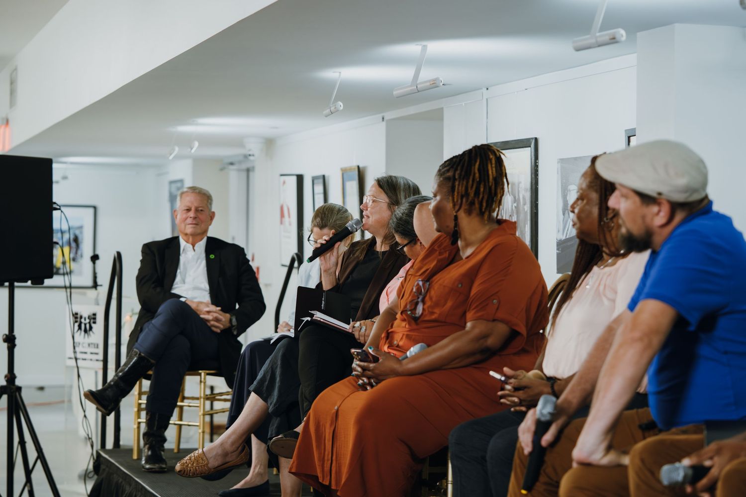 A wide panel discussion featuring seven diverse speakers seated in a row at a community event.
