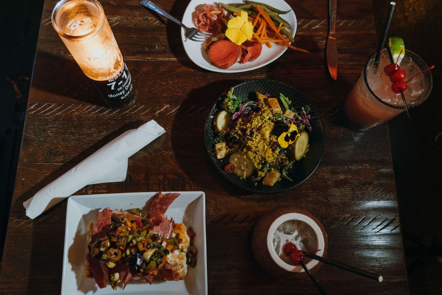Overhead view of a restaurant table with colorful salads, appetizers, and cocktails served on a dark wooden surface.