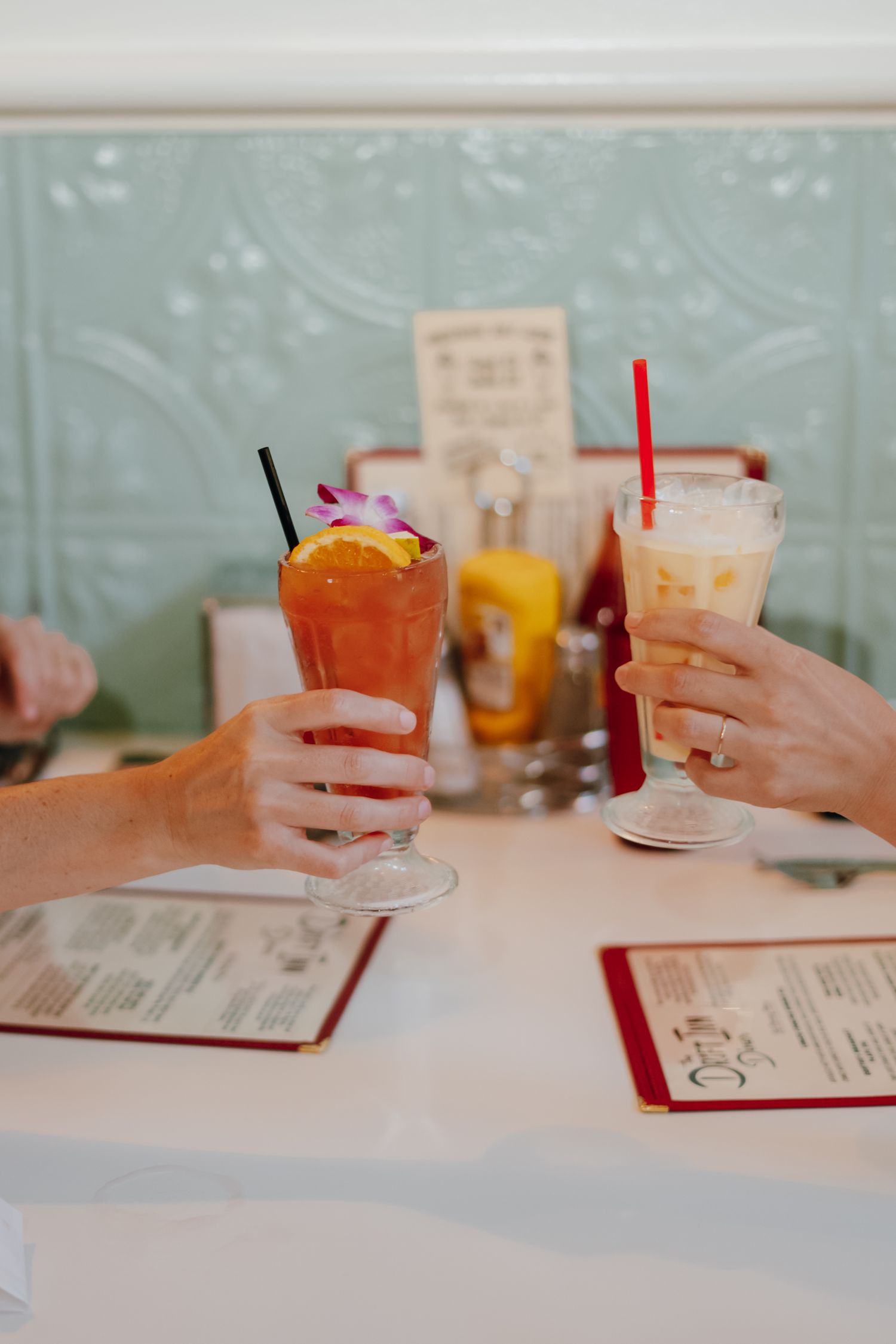 Two hands clinking colorful iced cocktails with straws at a retro diner table with menus.