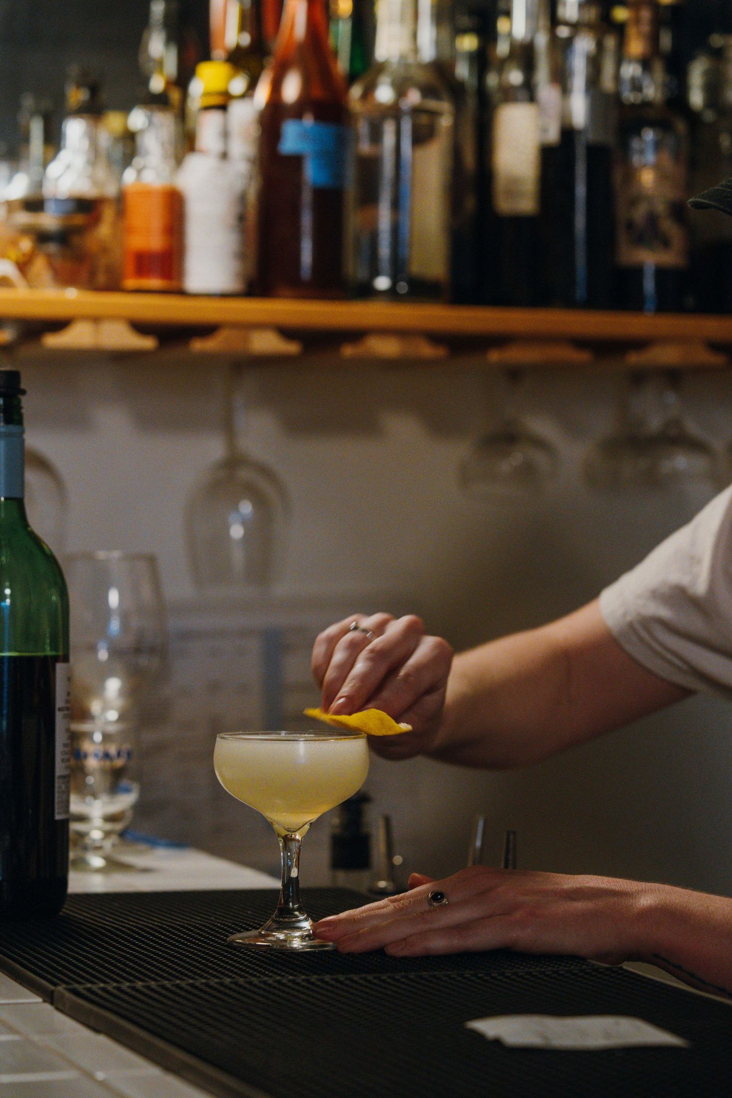 Bartender garnishing a yellow cocktail in a coupe glass at a bar with bottle-lined shelves.