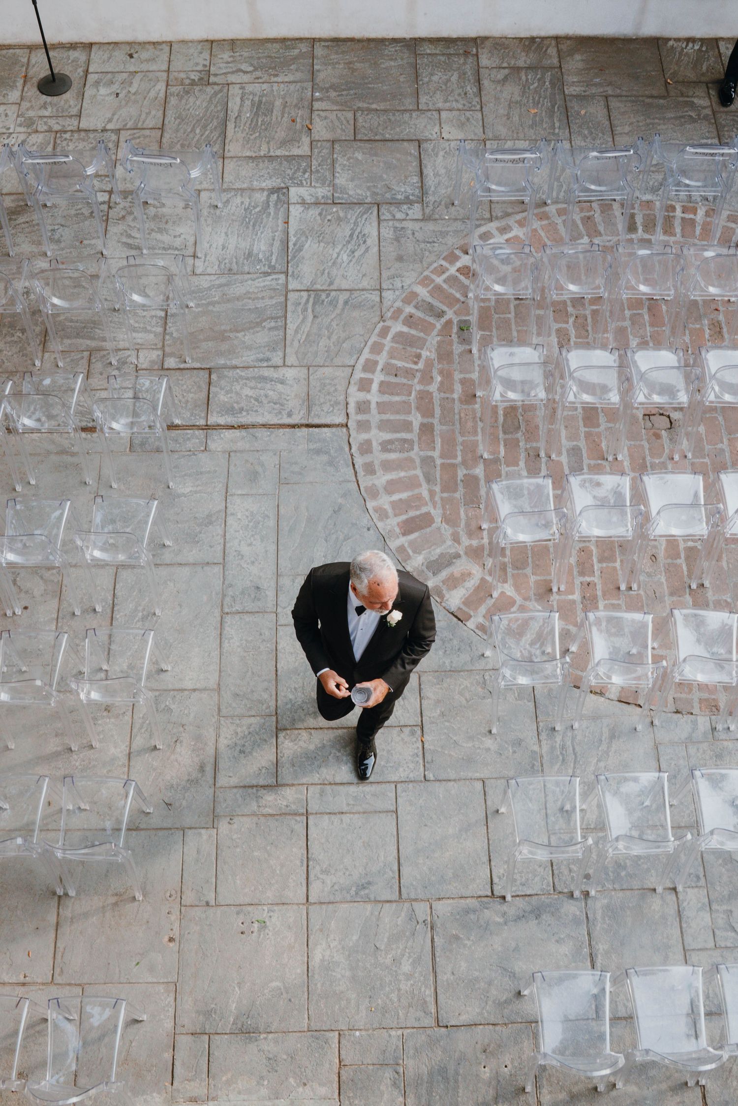 Aerial view of a man in a suit standing on a stone patio surrounded by clear acrylic chairs.