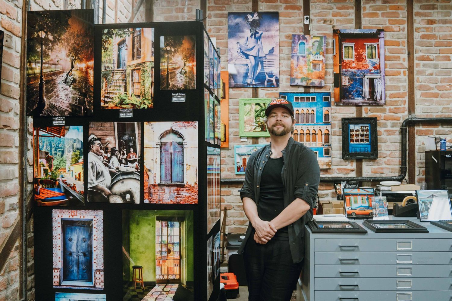 Man in black outfit and cap posing beside vibrant architectural photography prints on display.