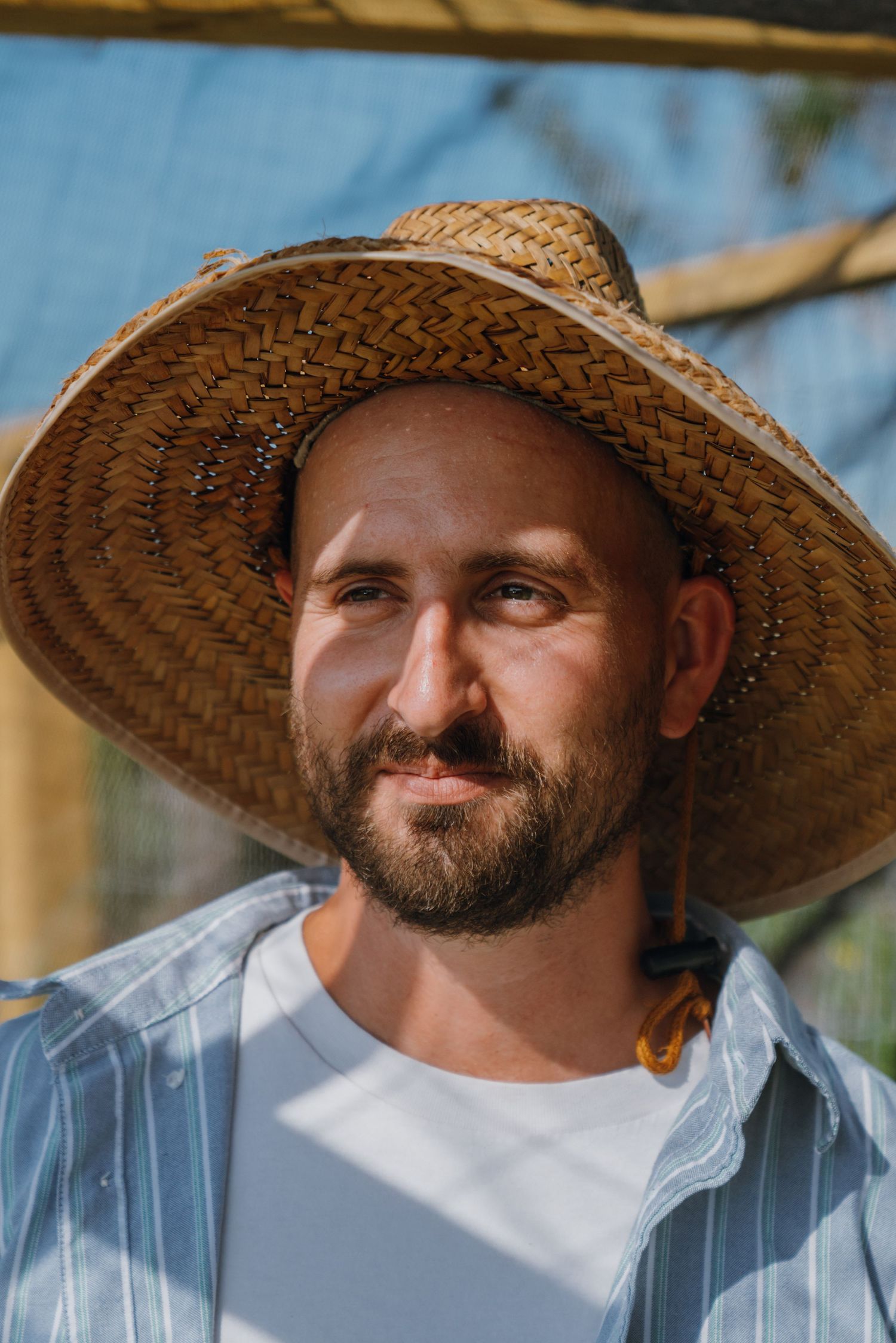 Bearded man wearing a woven straw hat smiling in bright sunlight outdoors.