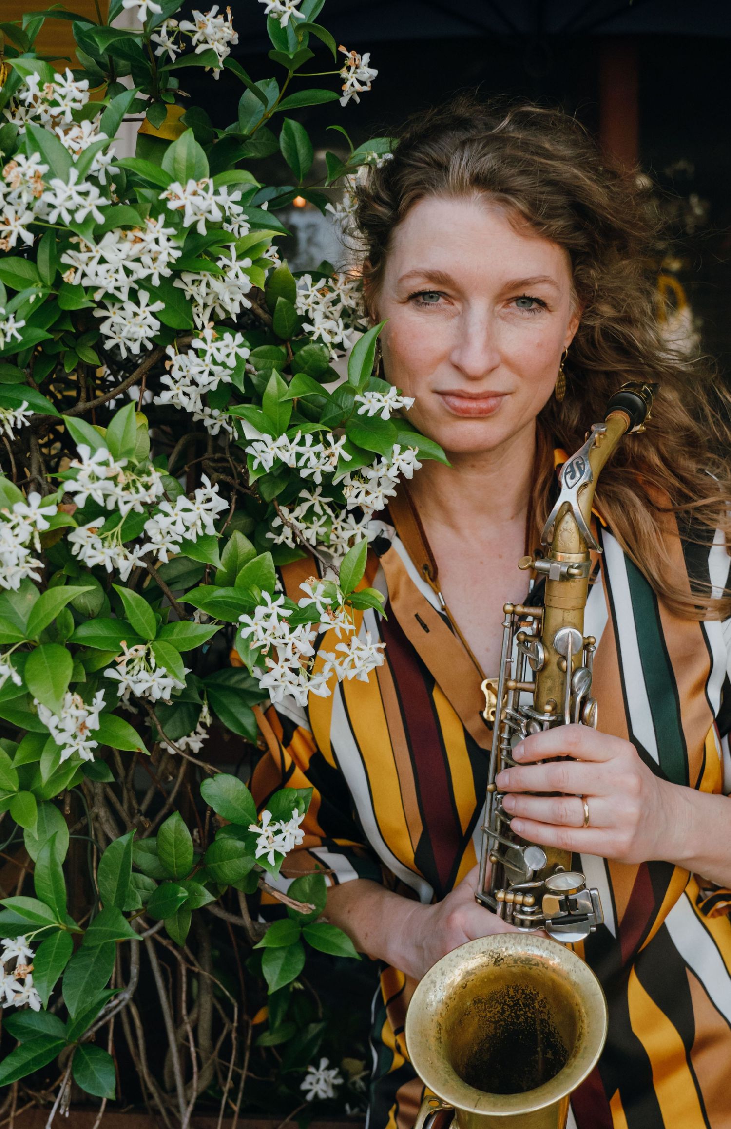 Woman holding a saxophone poses beside white flowering jasmine in a striped shirt outdoors.