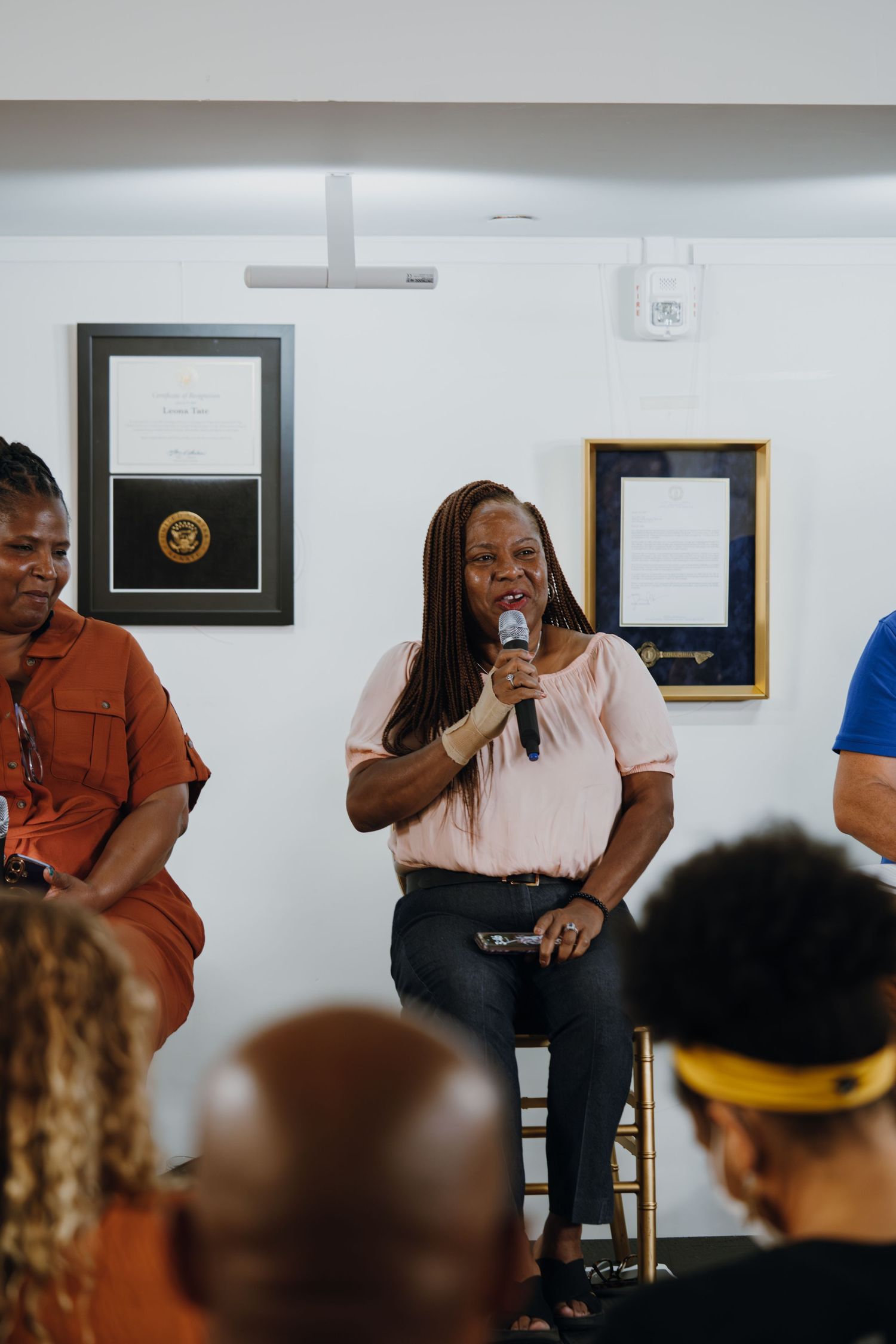 A Black woman speaks into a microphone during a panel discussion with framed certificates on the white wall behind her.