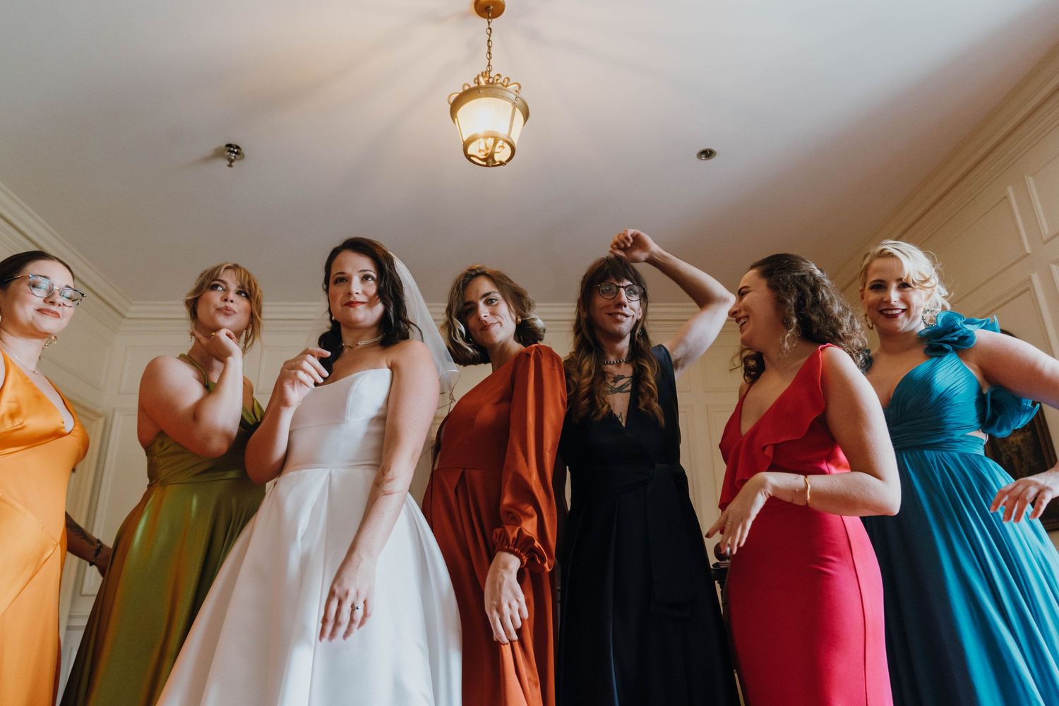 Bride in white gown laughing with bridesmaids in colorful dresses under a gold chandelier. New Orleans, Louisiana, Mississippi