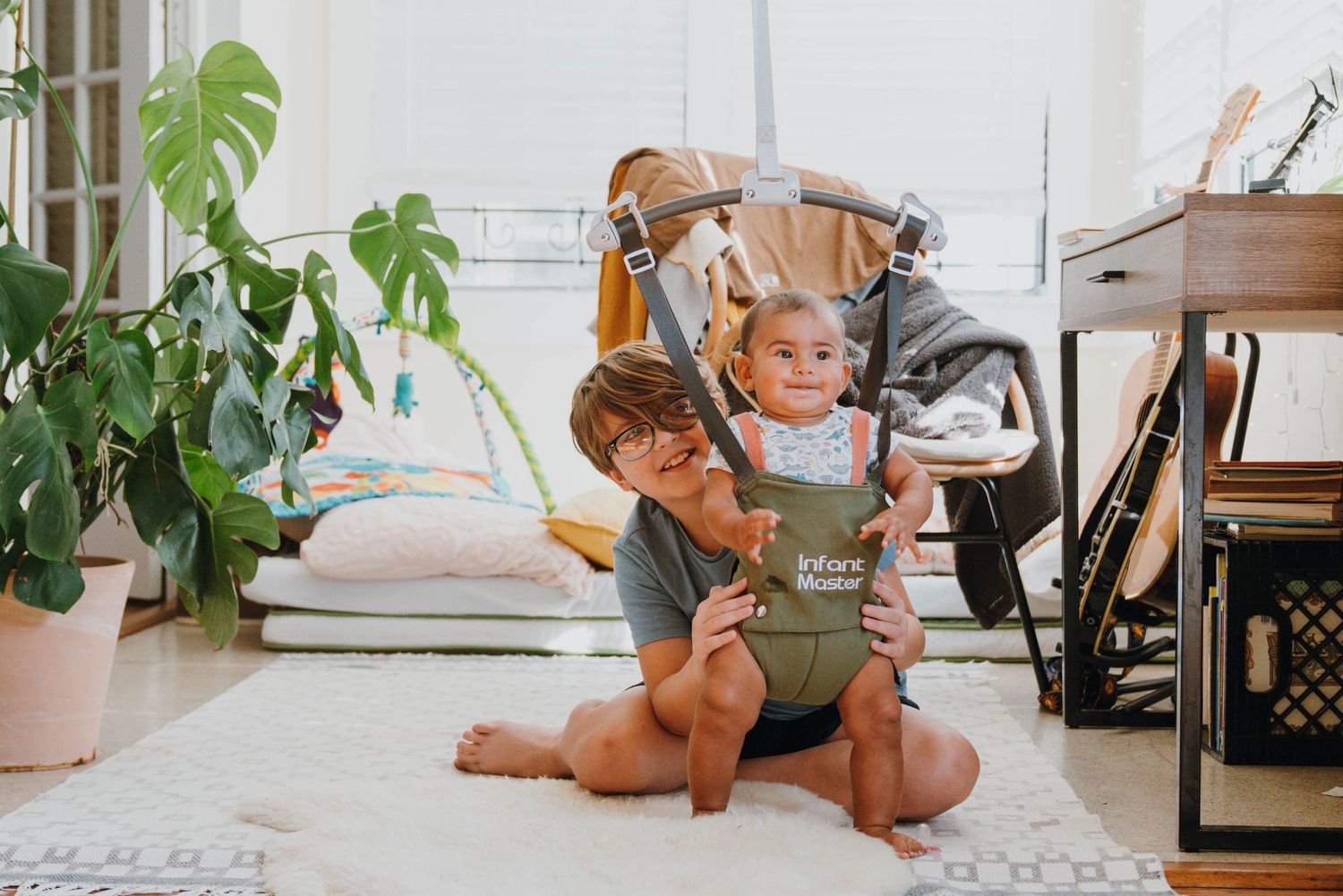 Parent sitting cross-legged on floor using baby carrier with smiling infant in bright airy living room.