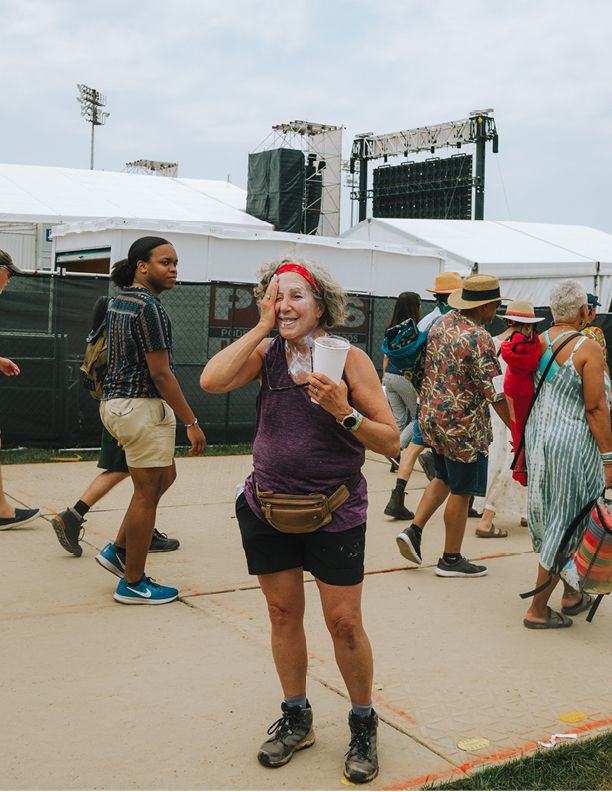 Woman smiling and holding a drink at an outdoor music festival with a stage and crowd in the background.