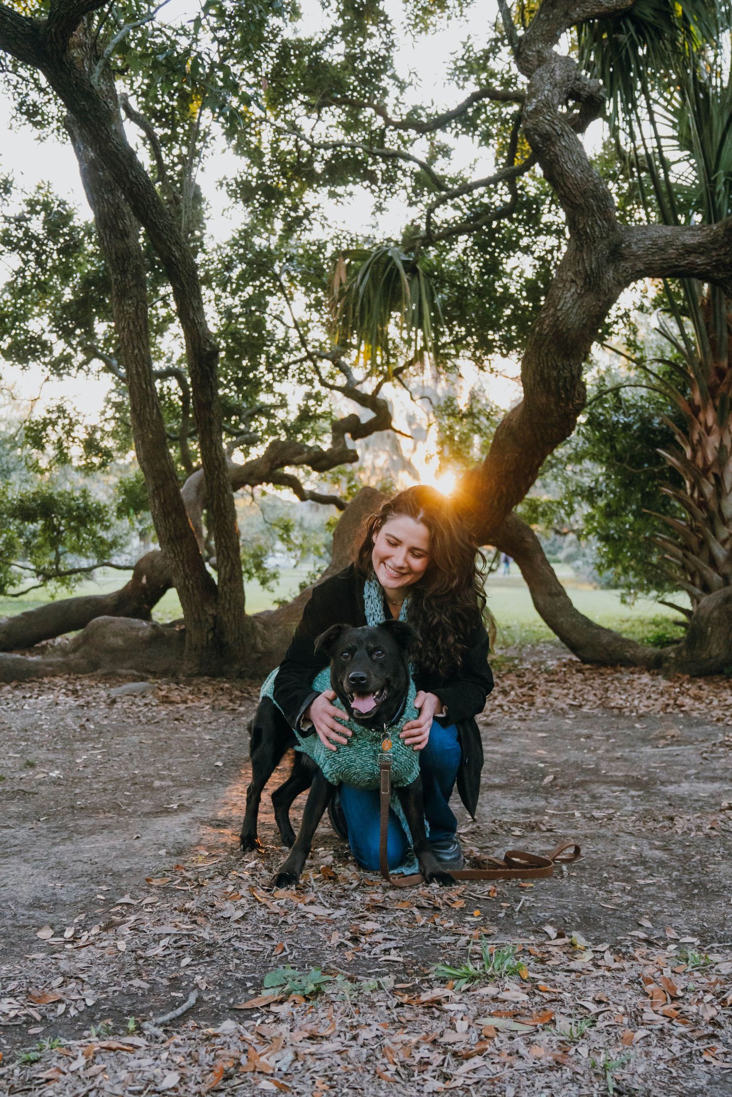 Smiling woman crouching with black dog wearing teal vest under sunlit trees in park.