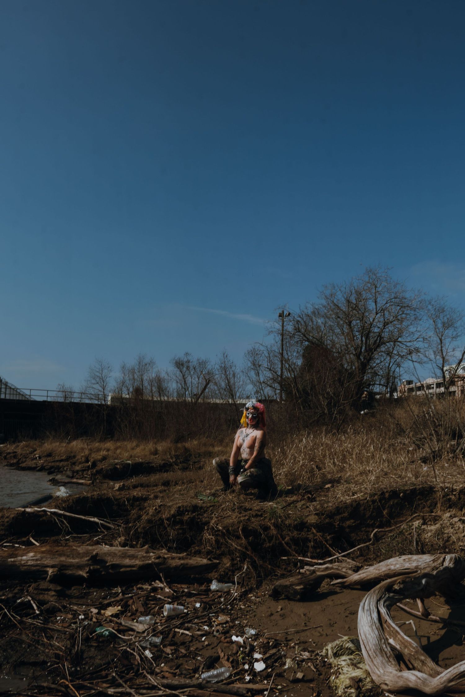 Person standing on a rocky riverbank surrounded by bare trees and driftwood under a clear blue sky.