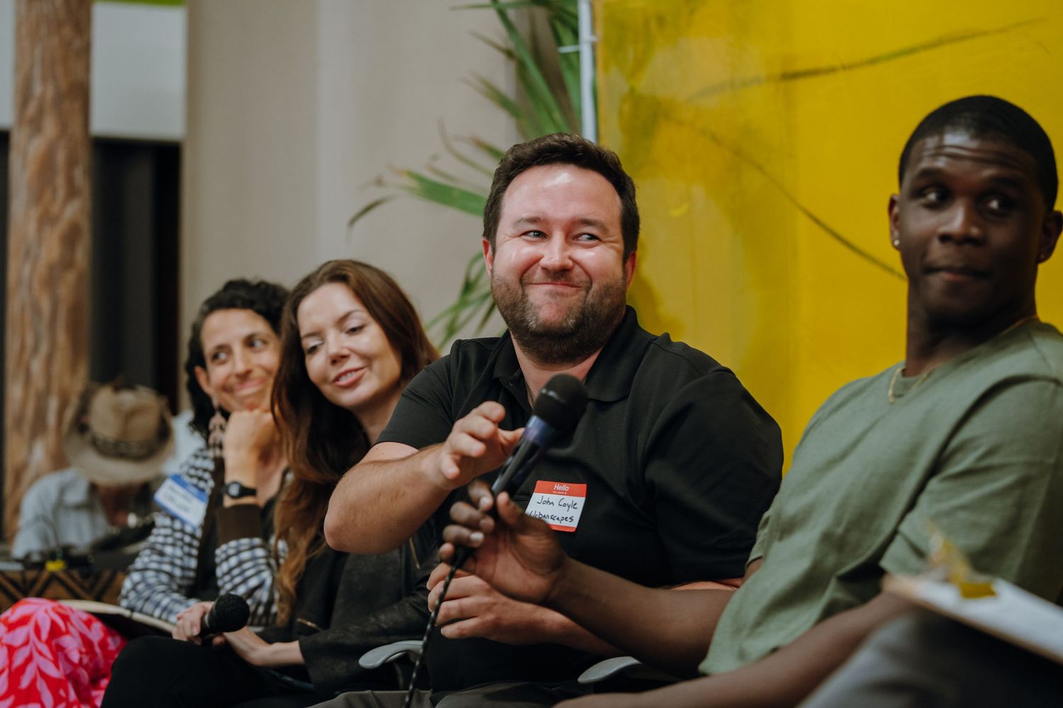 Panel discussion with diverse speakers smiling, man holding microphone against yellow backdrop.