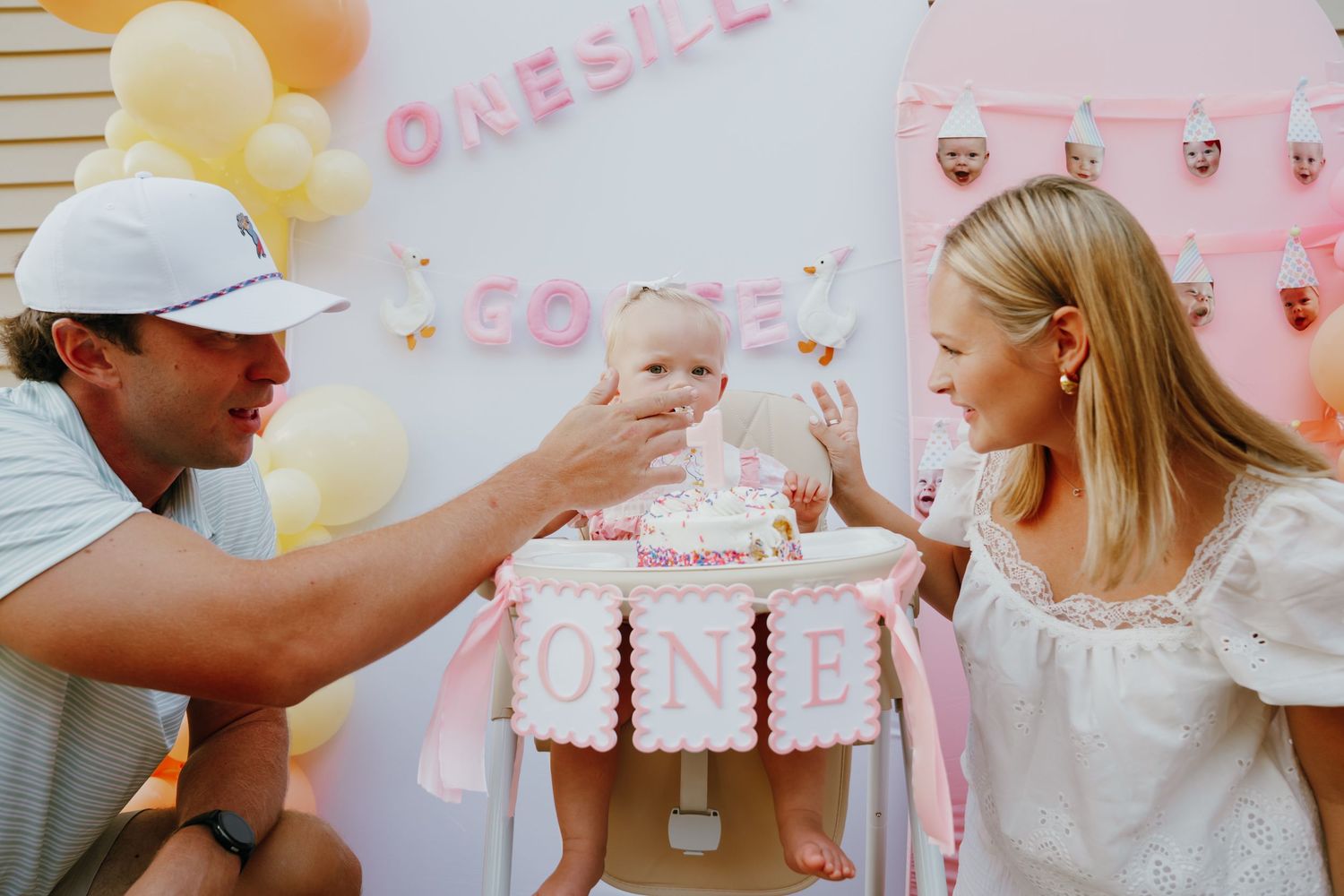 Parents celebrating baby's first birthday with a pink ONE cake at a decorated party.