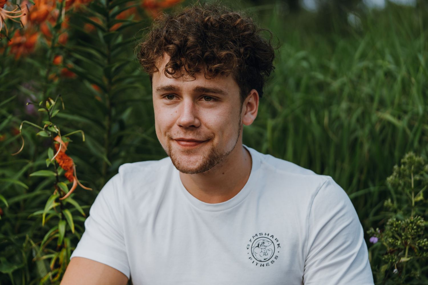 Young man with curly hair in white t-shirt smiles in front of lush green garden foliage.