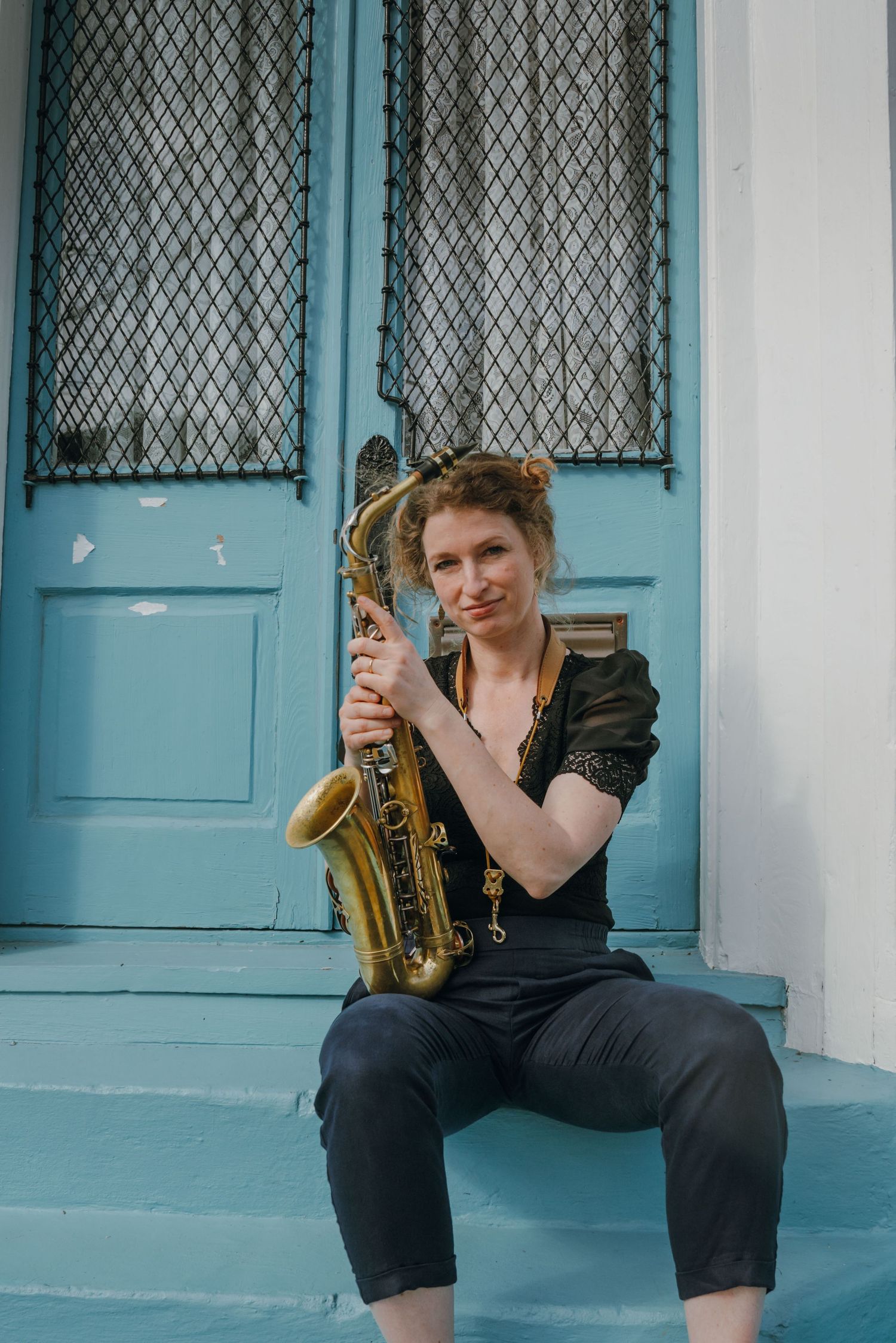 Female saxophonist sitting on blue porch steps holding brass saxophone in front of vintage door.