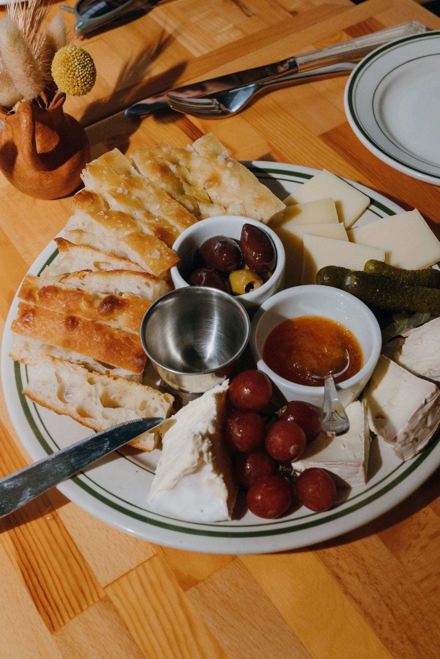 Close-up of a charcuterie board with bread, cheese, olives, jam, and pickles on a white plate.