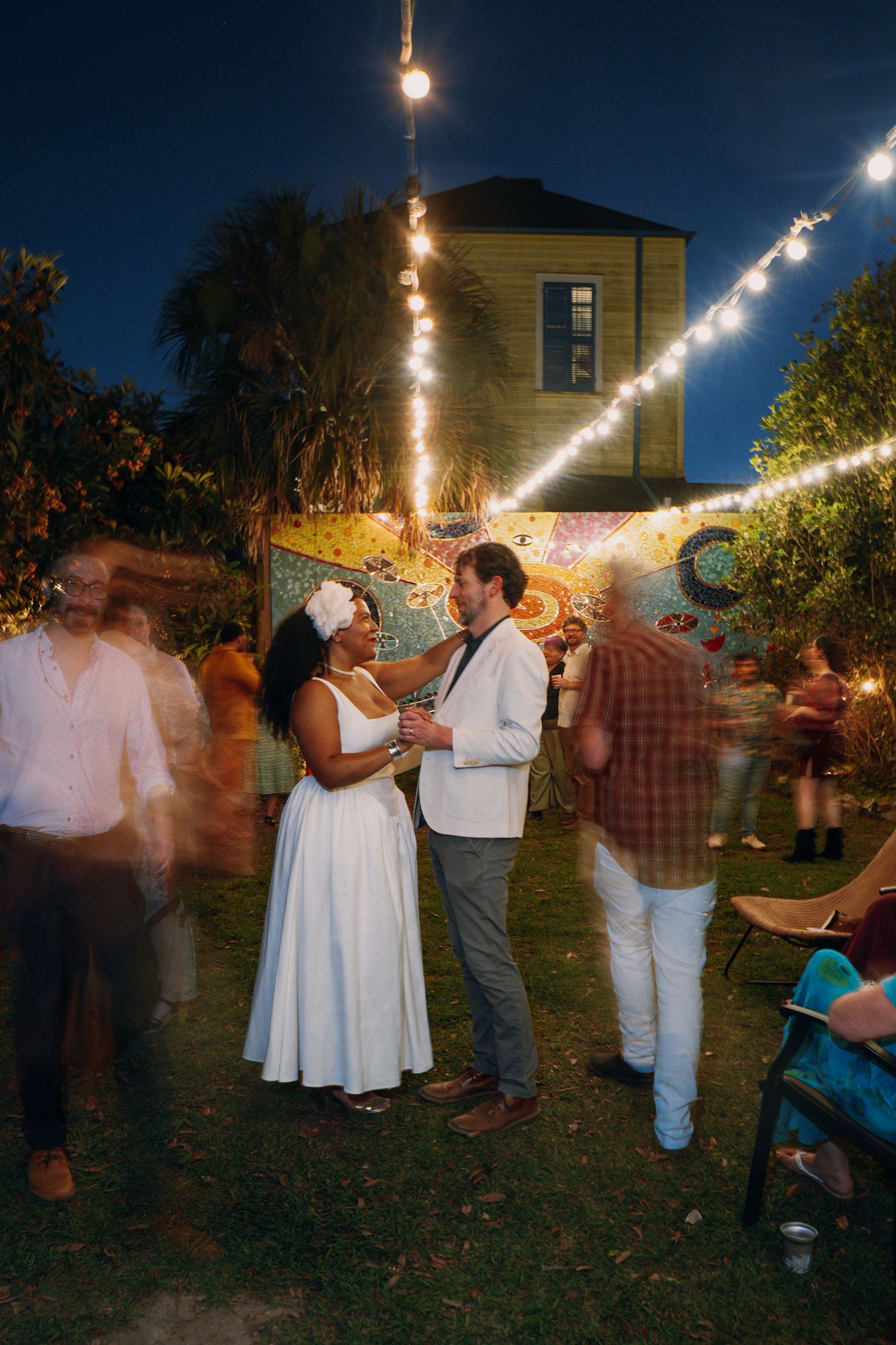 Bride and groom share a moment surrounded by dancing guests under string lights at outdoor wedding reception.