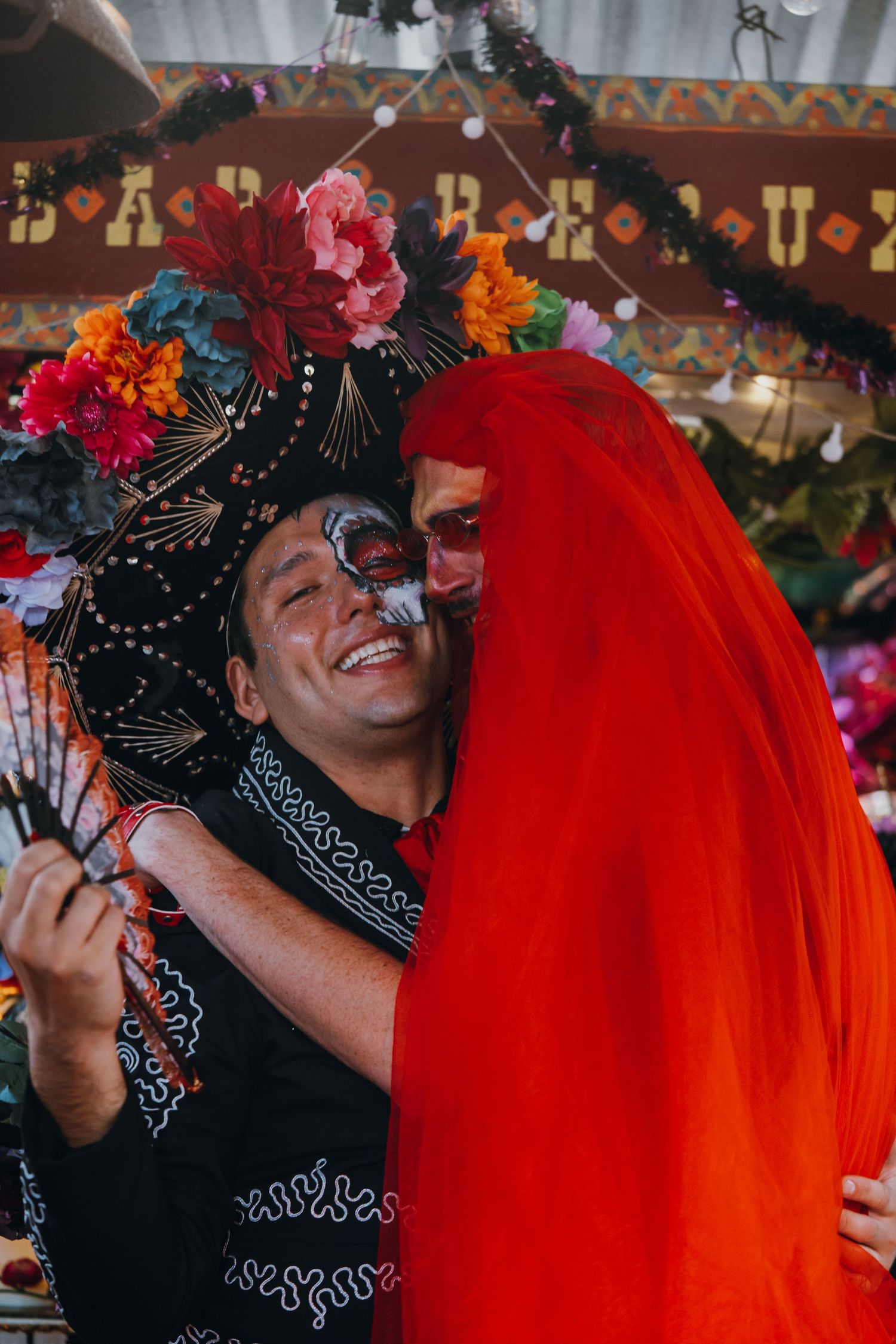 Two people in Day of the Dead face paint and costumes embrace and smile at a festive celebration.