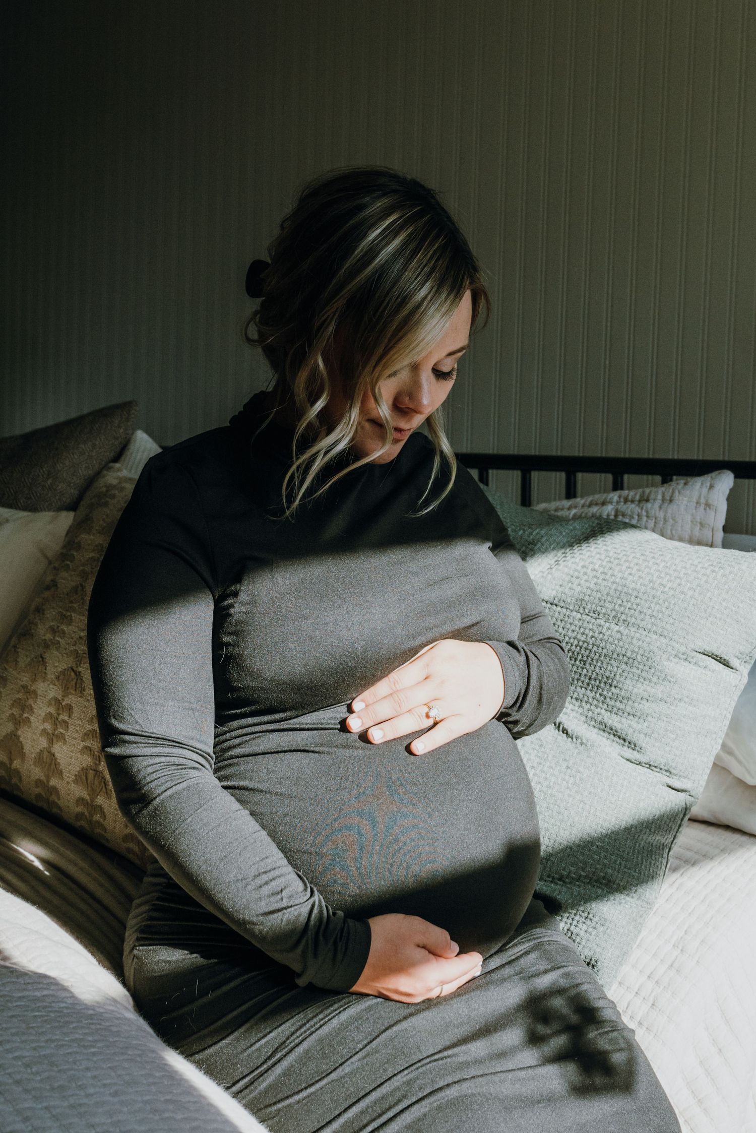Pregnant woman in black dress sitting on bed, tenderly holding her baby bump.