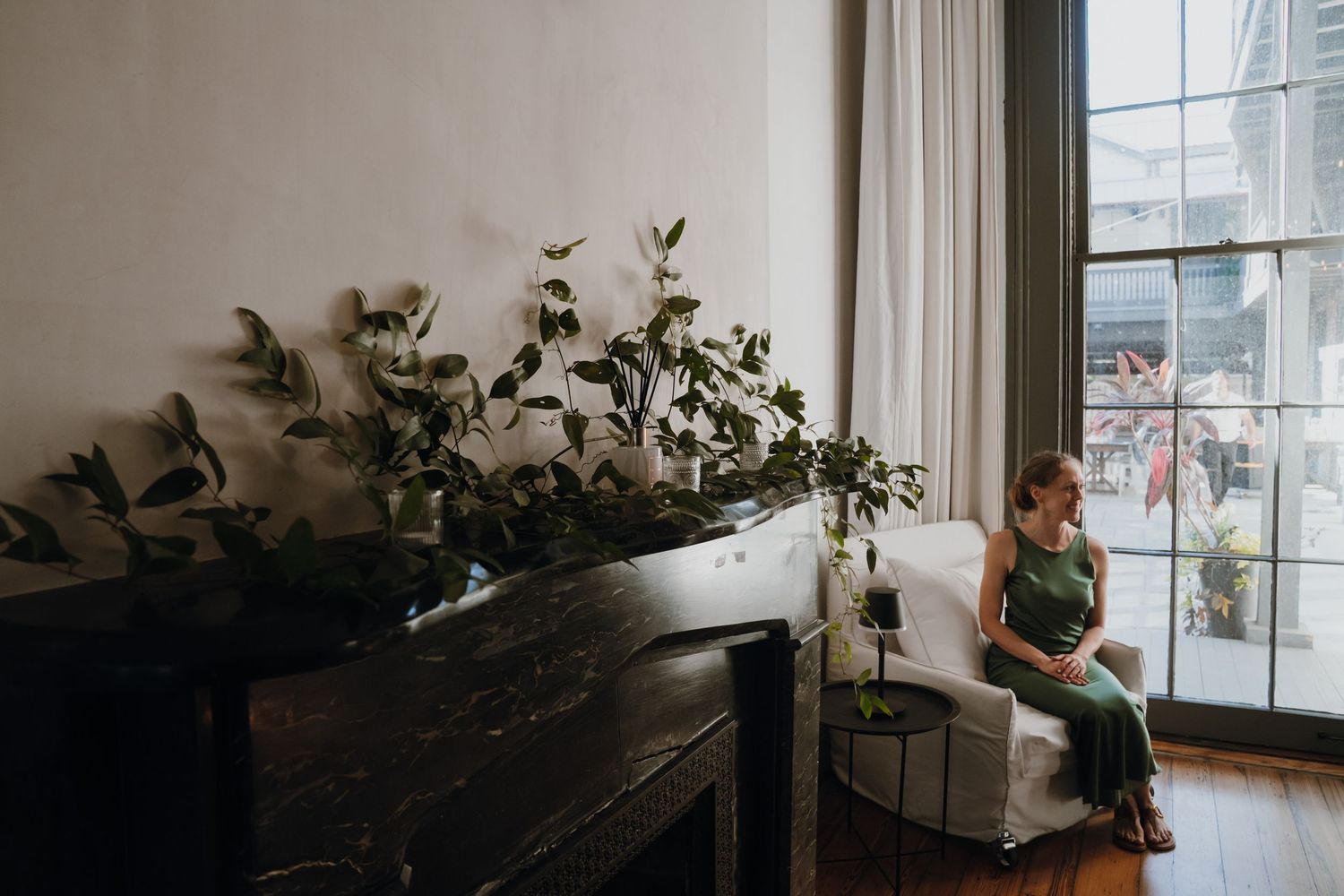 Woman in green dress sitting by large window near black piano with lush plants.