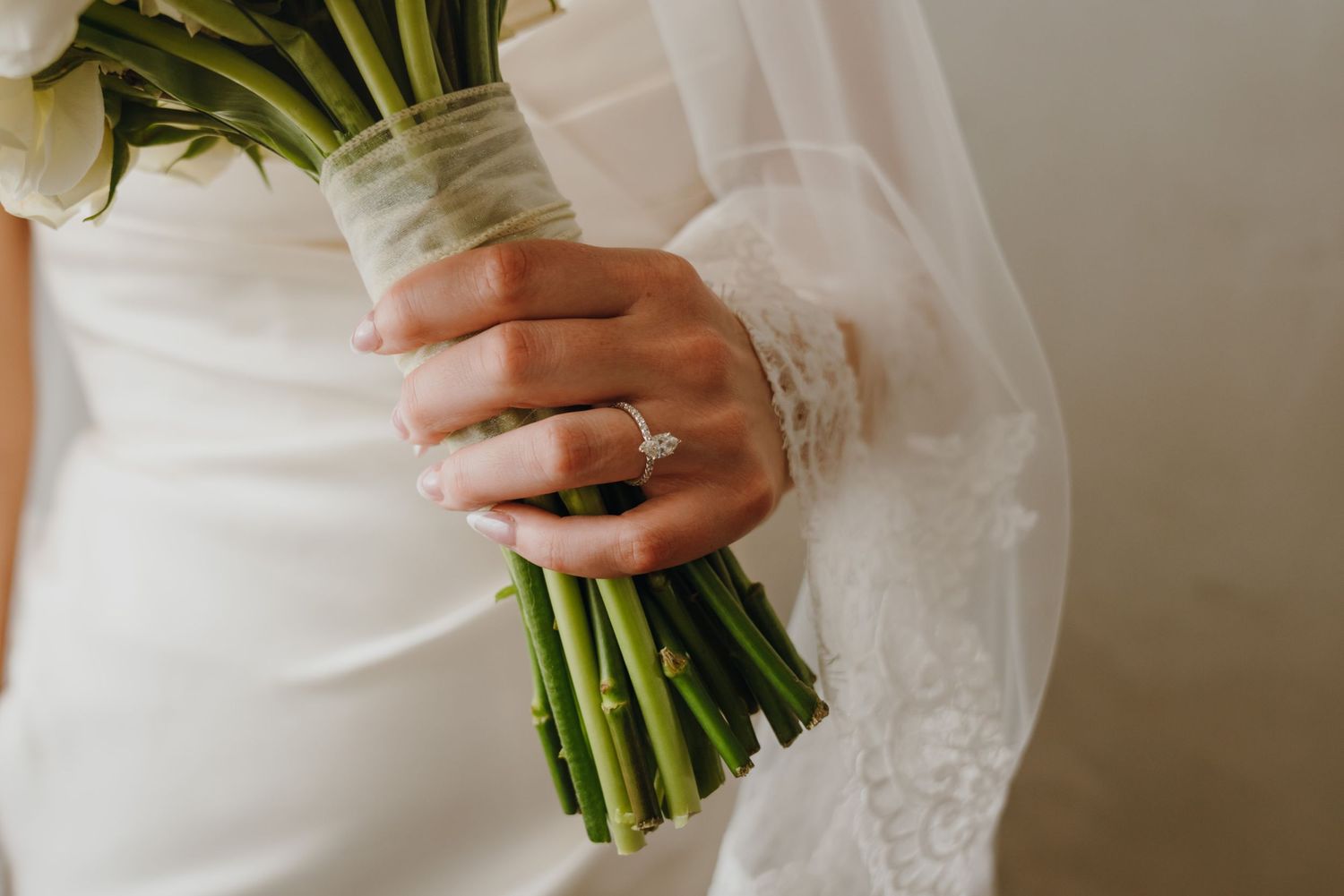 Close-up of bride holding white flower bouquet with diamond engagement ring on her finger in a white wedding dress.