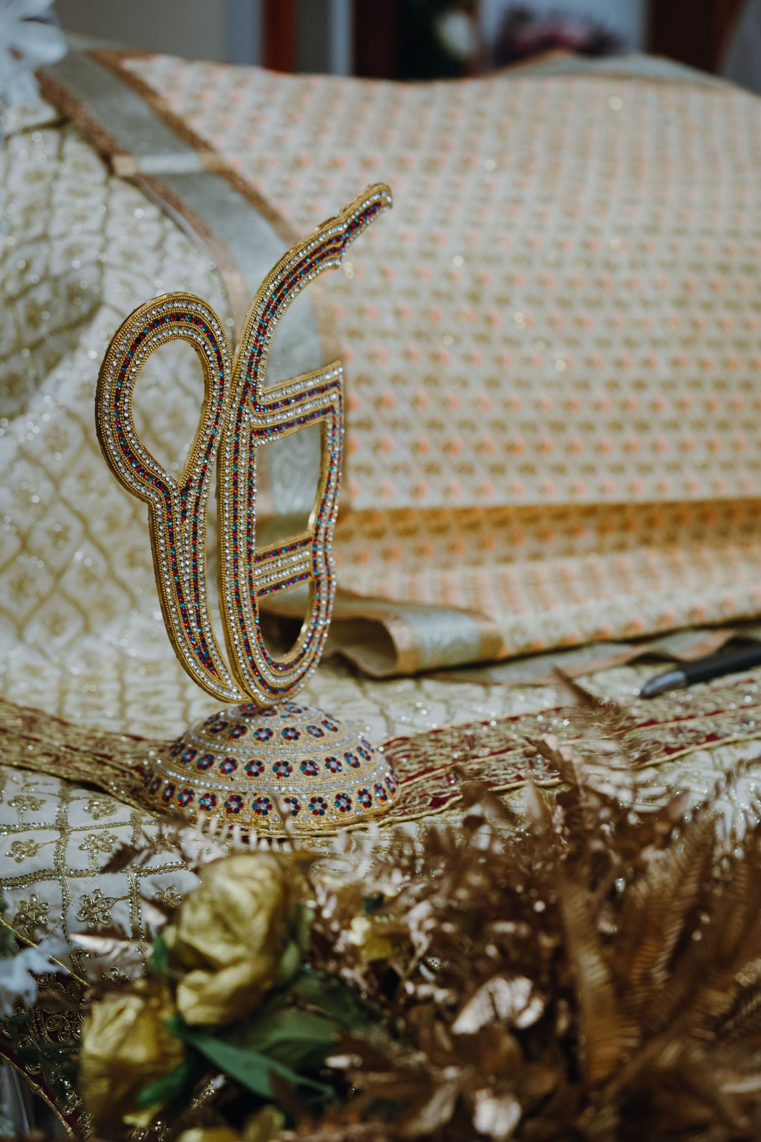 Ornate gold Ik Onkar symbol decorated with gemstones displayed among traditional Sikh wedding accessories.