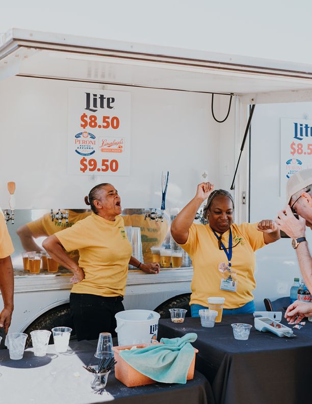 Two women in yellow shirts dance behind a Miller Lite beer stand at an outdoor event with prices $8.50 and $9.50.