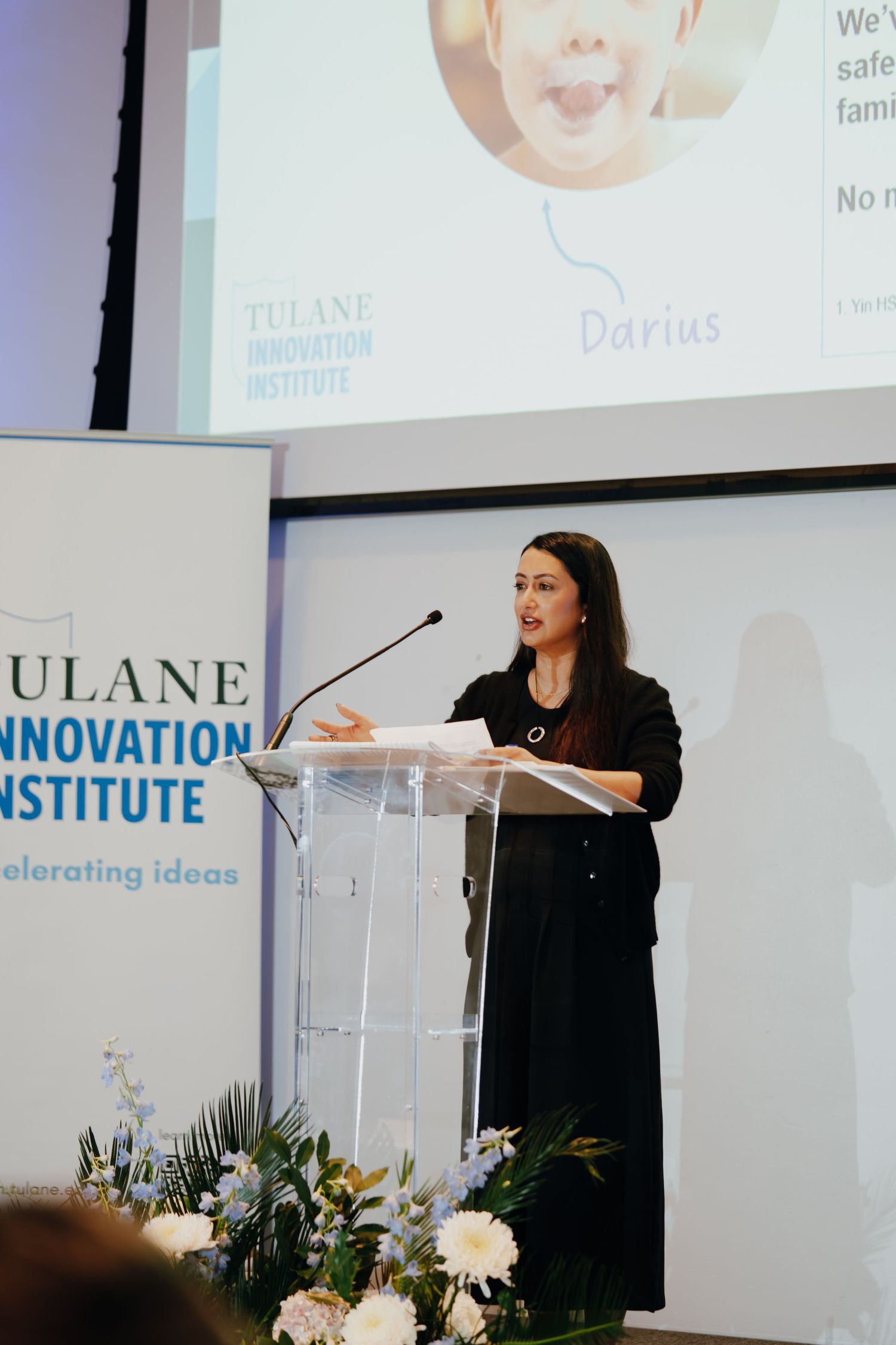 Woman presenting at Tulane Innovation Institute podium with projection screen displaying name Darius at conference event.