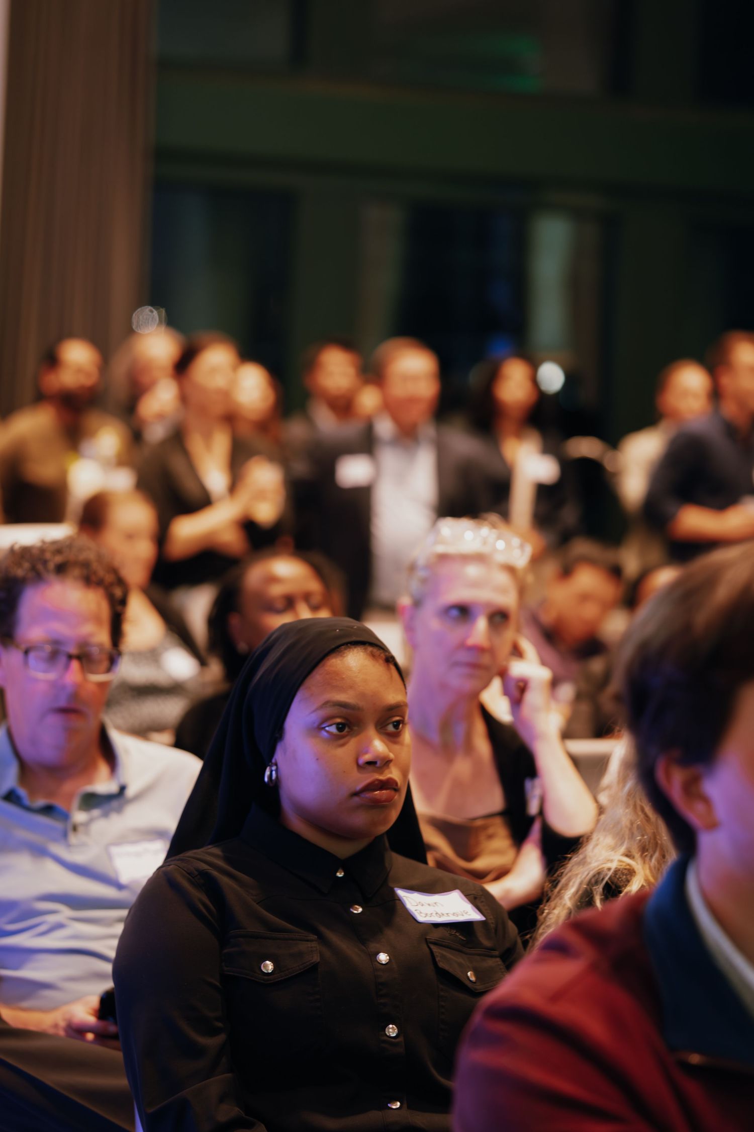 Diverse audience attends a professional networking event, with a woman in black headscarf in focus.