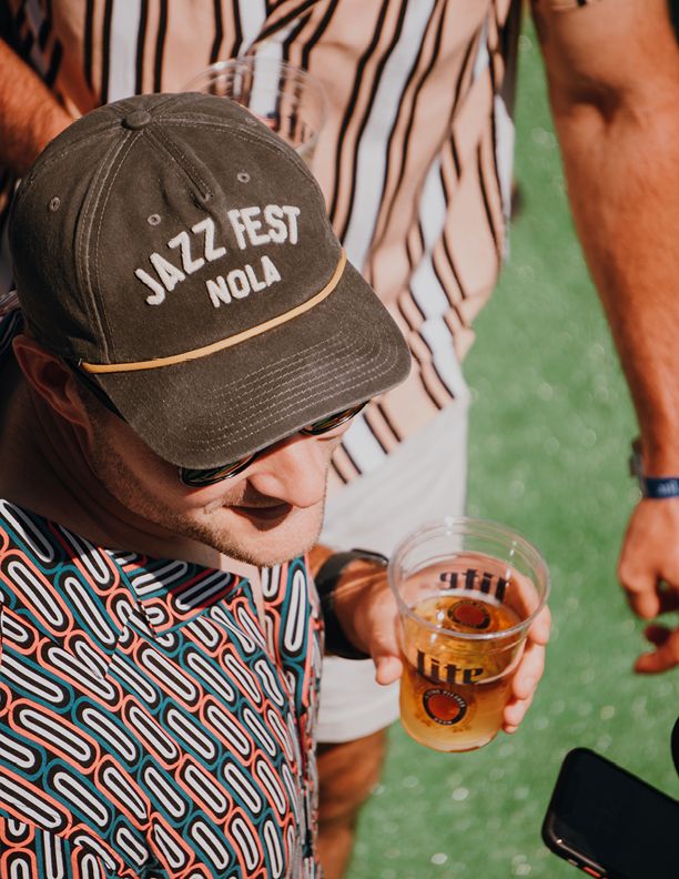 Man wearing Jazz Fest NOLA cap holding a beer at New Orleans Jazz Festival outdoor event.
