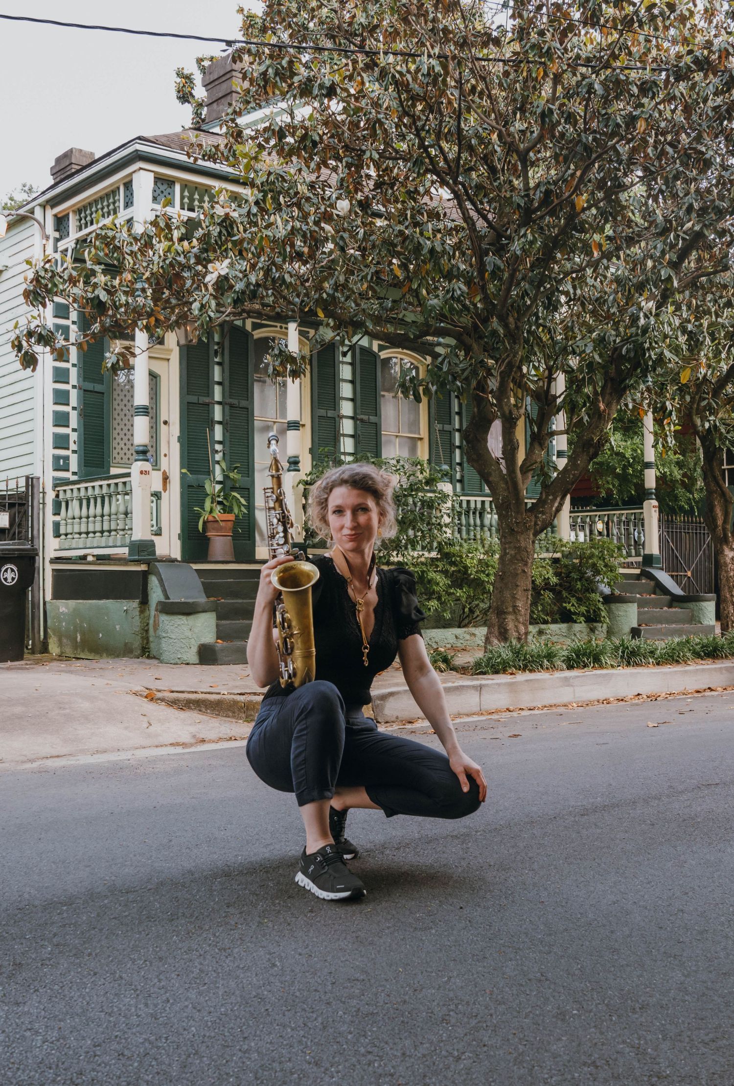 Woman crouching in street holding saxophone with historic New Orleans shotgun house in background.