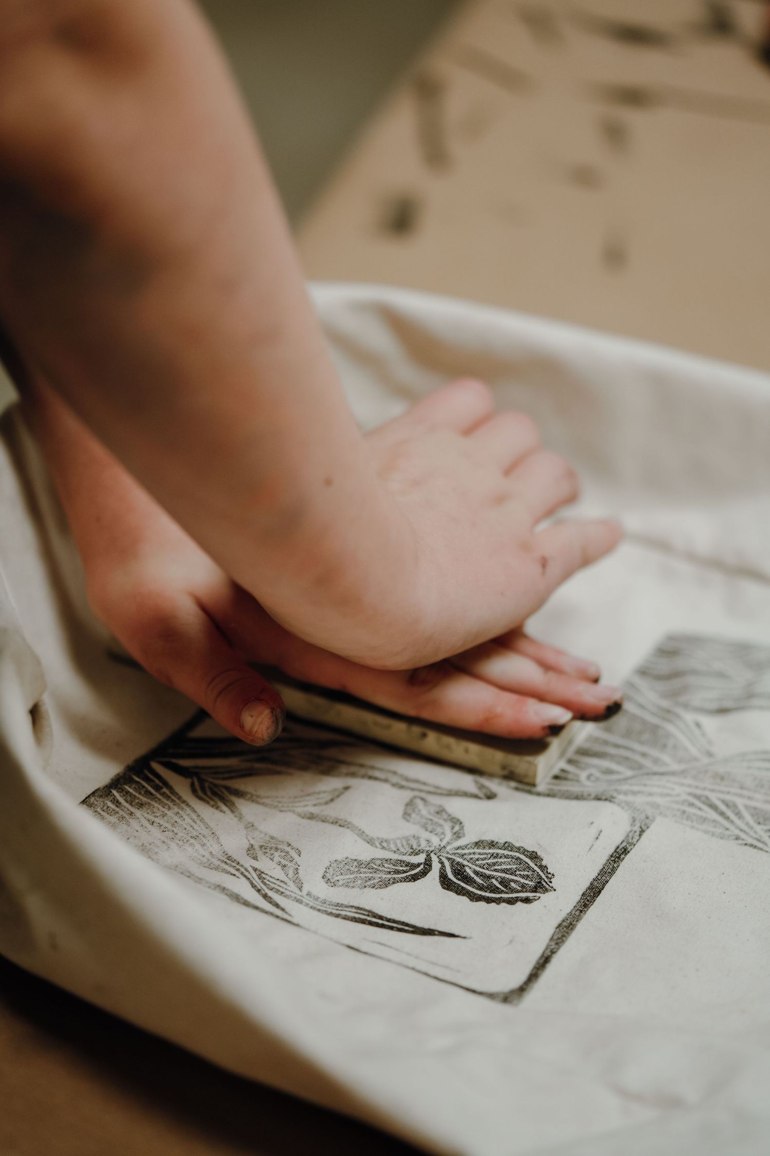 Hands pressing a botanical linocut block onto white fabric during a printmaking workshop.